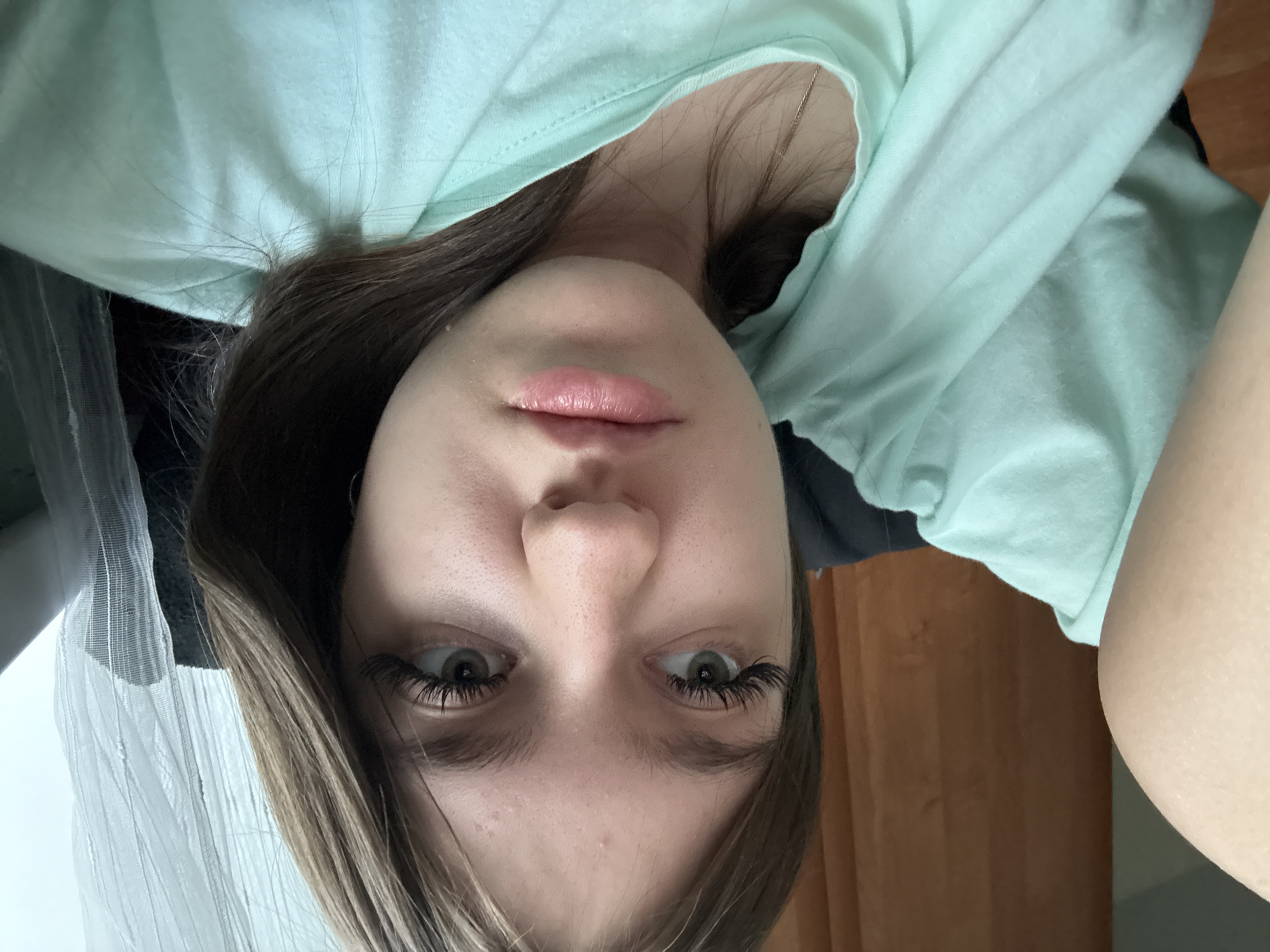 A close-up, high-angle portrait captures a young person with long brown hair, looking directly at the camera. The person is likely lying down, with their head positioned towards the lower edge of the frame. Their gaze is direct and neutral, with wide, light-colored eyes accentuated by long, dark eyelashes, which appear to be false or heavily mascaraed. The lips are slightly parted, revealing a soft pink color.
The individual is wearing a light mint green long-sleeved top. A thin, gold-colored chain is subtly visible around their neck in some of the cropped views. The soft, natural lighting suggests it is daytime.
In the background, on the left side, a sheer white curtain or fabric is visible, indicating the presence of a window. On the right side, a warm brown surface with a vertical wood grain is present, possibly a wall panel or furniture. No specific activity beyond posing for the camera is discernible. There is no visible text in the image.