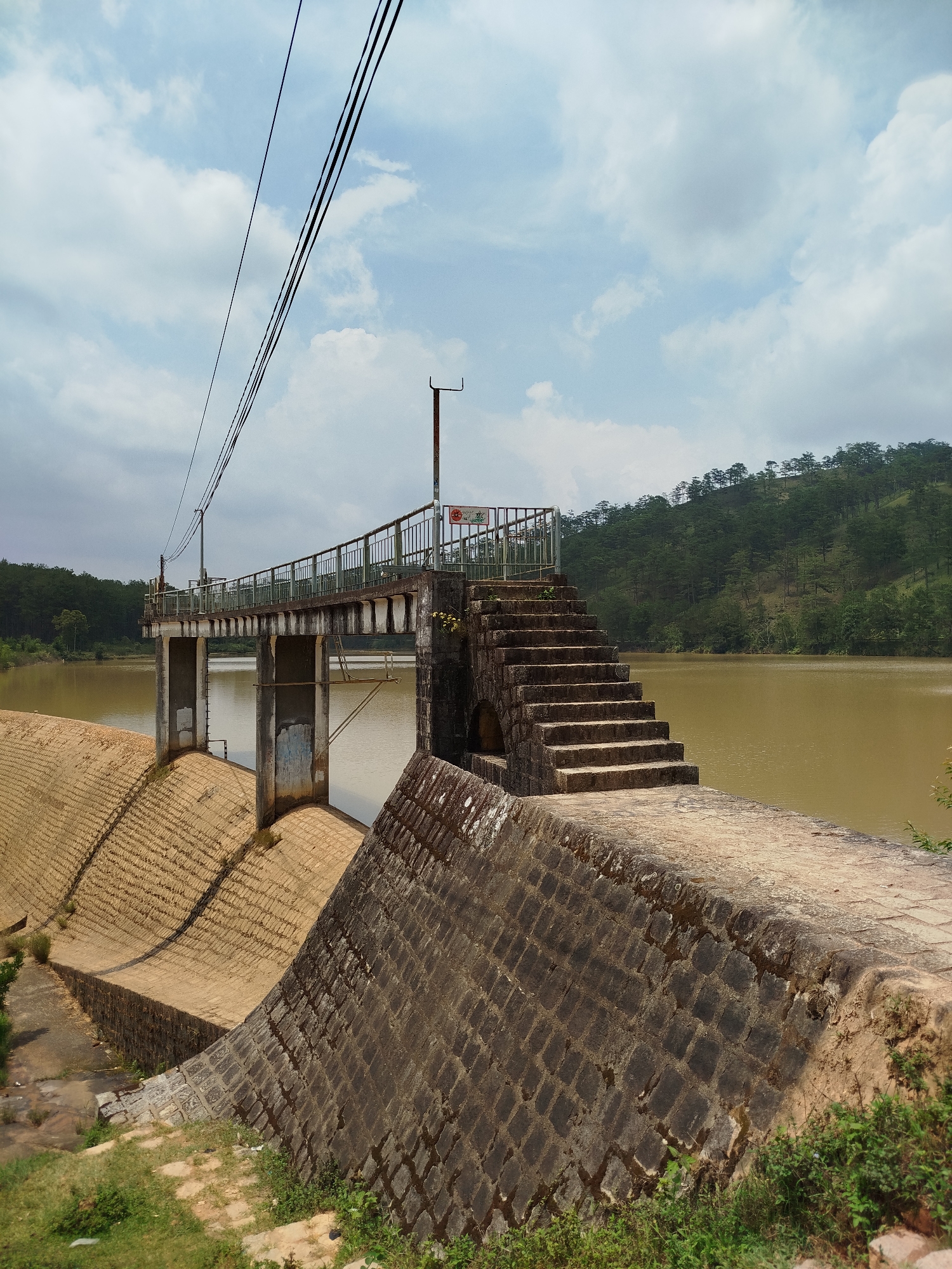 A daytime outdoor shot captures a dam structure and reservoir in Lang Biang, Da Lat, Vietnam, under a partly cloudy sky. The scene is devoid of people, highlighting the engineering marvel integrated into the natural landscape.
In the foreground, a textured stone wall, built with irregular blocks, forms part of the dam's lower section on the right, partially covered by moss and low green vegetation. This wall extends towards the left, where another section of the dam, a curved concrete structure with a patterned surface, is visible, suggesting a spillway or support. A set of weathered concrete stairs with stone risers ascends from the foreground wall to a higher platform on the right.
The midground features a functional concrete bridge with metal railings, supported by several concrete pillars over the murky, greenish-brown water of the reservoir. Power lines stretch across the top left of the frame, connecting to a utility pole located on the bridge. A small white sign with red and green text, largely unreadable, is attached to the bridge railing on the right side.
The background reveals a gently sloping hillside thickly covered with tall, slender evergreen trees, likely pines, characteristic of the Da Lat region. The sky above is a mix of pale blue and scattered white and grey clouds, indicating overcast or partly cloudy weather. The overall impression is one of a serene yet functional infrastructure within a natural setting.