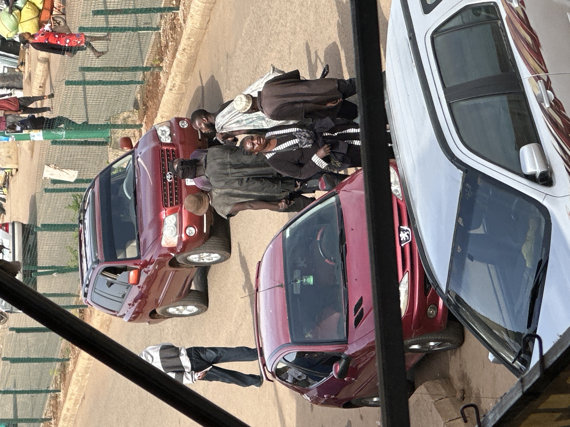 In Bukuru, Nigeria, a street scene unfolds. A red Toyota Highlander SUV is parked on the right side of the road. A group of people are gathered near the SUV, some appearing to be in conversation. One man, wearing a brown hat and a grey tunic, stands next to the SUV. Behind him, another man in a dark jacket and a checkered shirt is visible. A woman in a black outfit with white trim is in the foreground, looking towards the left. Another man in a brown long-sleeved shirt and a light-colored cap is standing to her right. A red hatchback car is parked in front of the Highlander. In the background, a white SUV is partially visible. The street is paved and a wire fence runs along the side of the road, with some vegetation behind it. A person in a black and white striped jacket and dark pants is walking away from the camera on the left side of the scene. Further in the background, more people and vehicles are present.