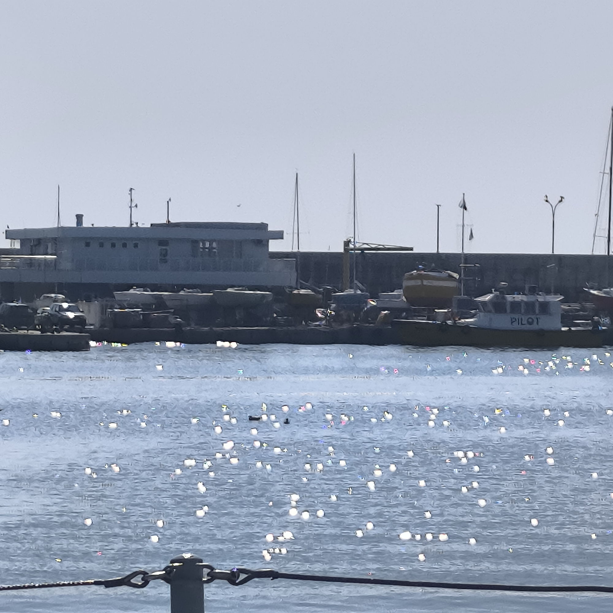 The image depicts a harbor scene in Varna, Bulgaria. In the foreground, a chain fence runs horizontally across the bottom, with a dark bollard visible to the right. The water in the middle ground is choppy, reflecting bright sunlight in numerous shimmering spots. Several small dark shapes, possibly ducks or debris, are scattered across the water's surface.
In the background, a harbor wall stretches across the width of the image. Moored to the wall are various vessels. Most prominently, a white boat with "PILOT" written on its side is visible. To its left, a large yellow cylindrical object, likely a buoy, is mounted on a boat. Further to the left, several other boats and structures are partially obscured. A building with a flat roof and numerous windows is situated behind the moored boats. Several masts from sailboats or other vessels are also visible, piercing the sky. The sky itself is a pale, hazy grey, suggesting an overcast or hazy day. The overall impression is one of a working harbor with a calm but bright atmosphere.