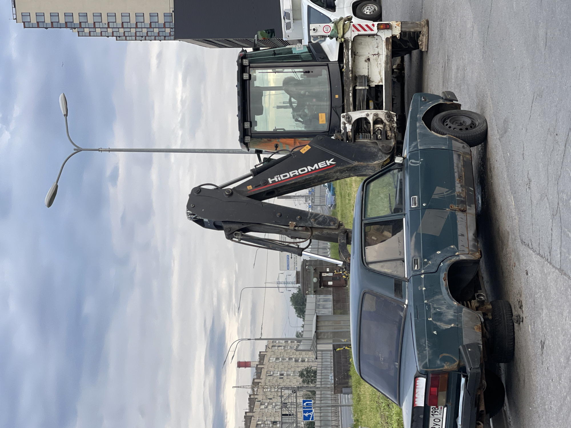 In an outdoor urban setting, a dark green, heavily rusted sedan sits on a cracked asphalt road, missing its rear tires. An excavator with the brand name "HIDROMEK" is positioned behind and to the right of the sedan. The excavator is white and black, with its arm extended. In the background, there are tall buildings, streetlights, and industrial structures under a cloudy sky. The car has a license plate that reads "2YO 198 RUS". There are several circular indentations on the side of the car's body.