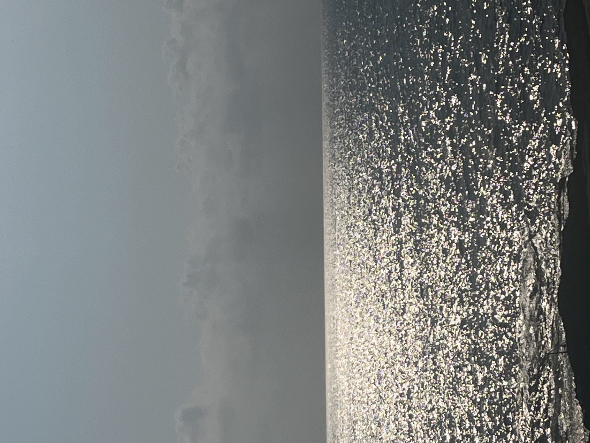 A wide-angle, eye-level shot captures a vast expanse of shimmering ocean under a hazy, overcast sky. The water's surface is a dynamic mosaic of light reflections, creating a sparkling effect as sunlight pierces through the clouds. The horizon line is sharp, dividing the textured water from the muted tones of the sky. In the foreground, a dark, rough edge of land, possibly a coastline or breakwater, juts into the frame from the bottom, its texture contrasting with the smooth water. The location is identified as the city of Cabo Cervera-Playa La Mata, Spain, though no specific landmarks or inhabitants are visible in this serene, naturalistic scene.