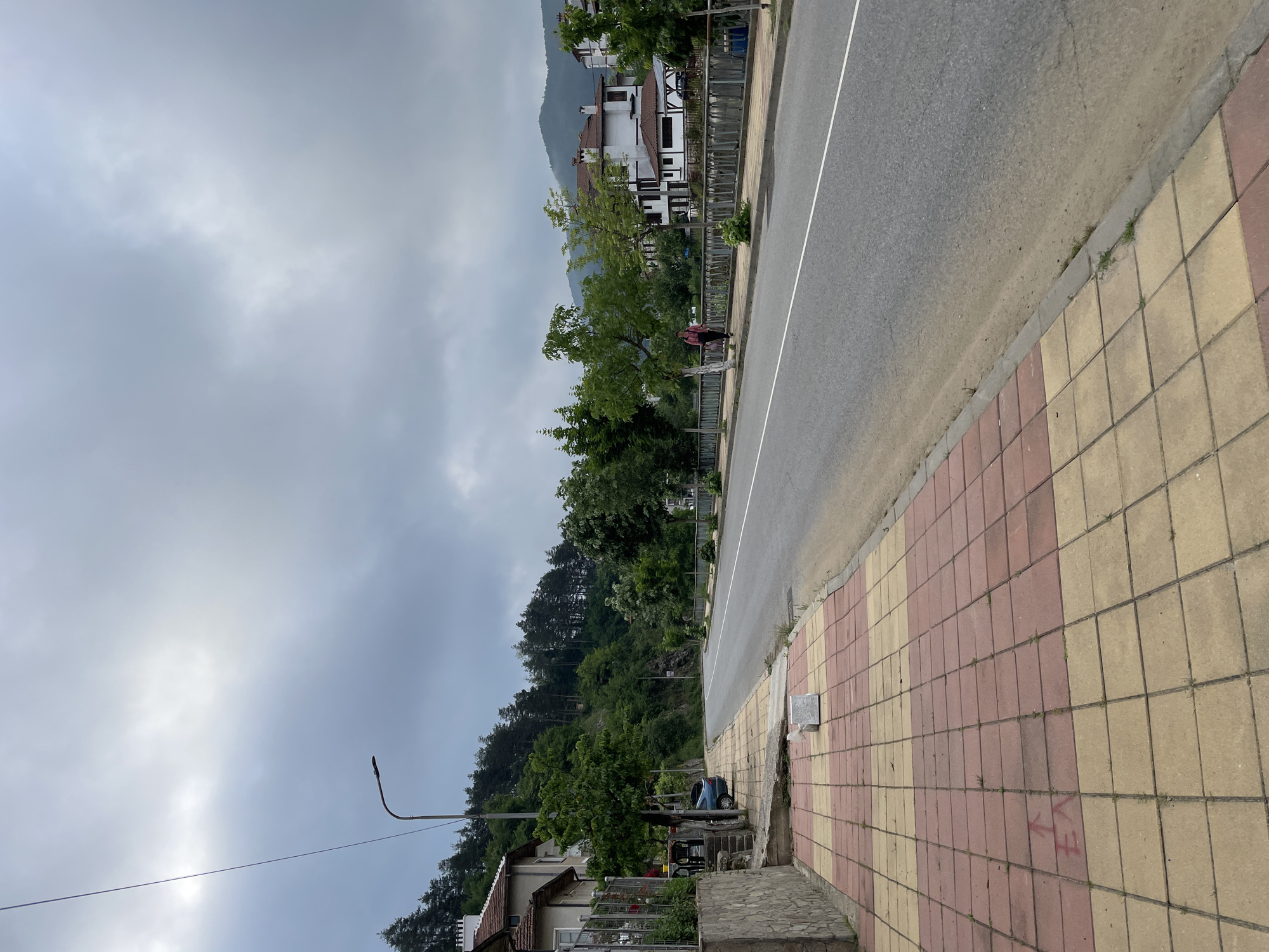 The image presents an overcast street scene in a mountainous town, likely Zlatograd, Bulgaria. The view looks along an asphalt road that gently curves, bordered by a tiled sidewalk on the right.
The sky is predominantly covered with light grey, thick clouds, suggesting an overcast day with diffuse lighting. Lush, dense green trees and foliage densely cover the steep hills and mountains that rise behind the visible structures, indicating a rich natural environment.
On the right side, a sidewalk constructed from alternating light yellow and reddish-brown rectangular tiles runs parallel to the road. A white solid line marks the edge of the asphalt. A person wearing dark clothing is walking away from the viewer on this sidewalk, further down the street. Adjacent to the road, a metal fence separates the public space from private properties. Behind this fence, several multi-story buildings with white facades, dark roofs, and some traditional half-timbered architectural details are visible, nestled against the mountainside.
On the left side, another segment of the tiled sidewalk is visible, featuring a black, curved street lamp and a power line stretching horizontally across the middle of the frame. Below the power line, a house with a tiled roof and a small garden is partially seen, with a small blue car parked nearby. Another individual is partially visible near this house, obscured by a tree.
On the tiled sidewalk in the lower right corner, there is red spray paint forming what appears to be the letters "EA" or "E/A" with an upward-pointing arrow. The scene conveys a calm, residential atmosphere in a scenic, hilly setting. The ground appears dry, and the time of day is clearly daytime, characterized by soft, indirect light.
