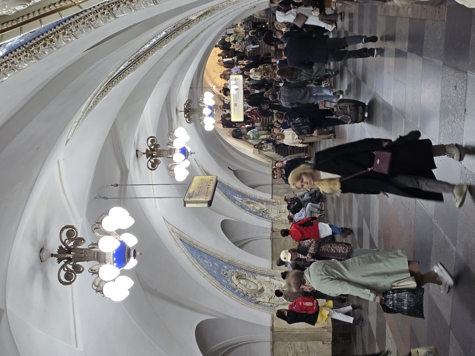 The image shows a busy underground scene, possibly a subway station, filled with people. The architecture is distinctive with a vaulted ceiling featuring layered, curved panels and ornate chandeliers. The walls have decorative panels with blue and white designs. People are walking in various directions, some with luggage, suggesting they may be commuters or travelers. The scene is well-lit, and the floor is tiled in a checkered pattern.