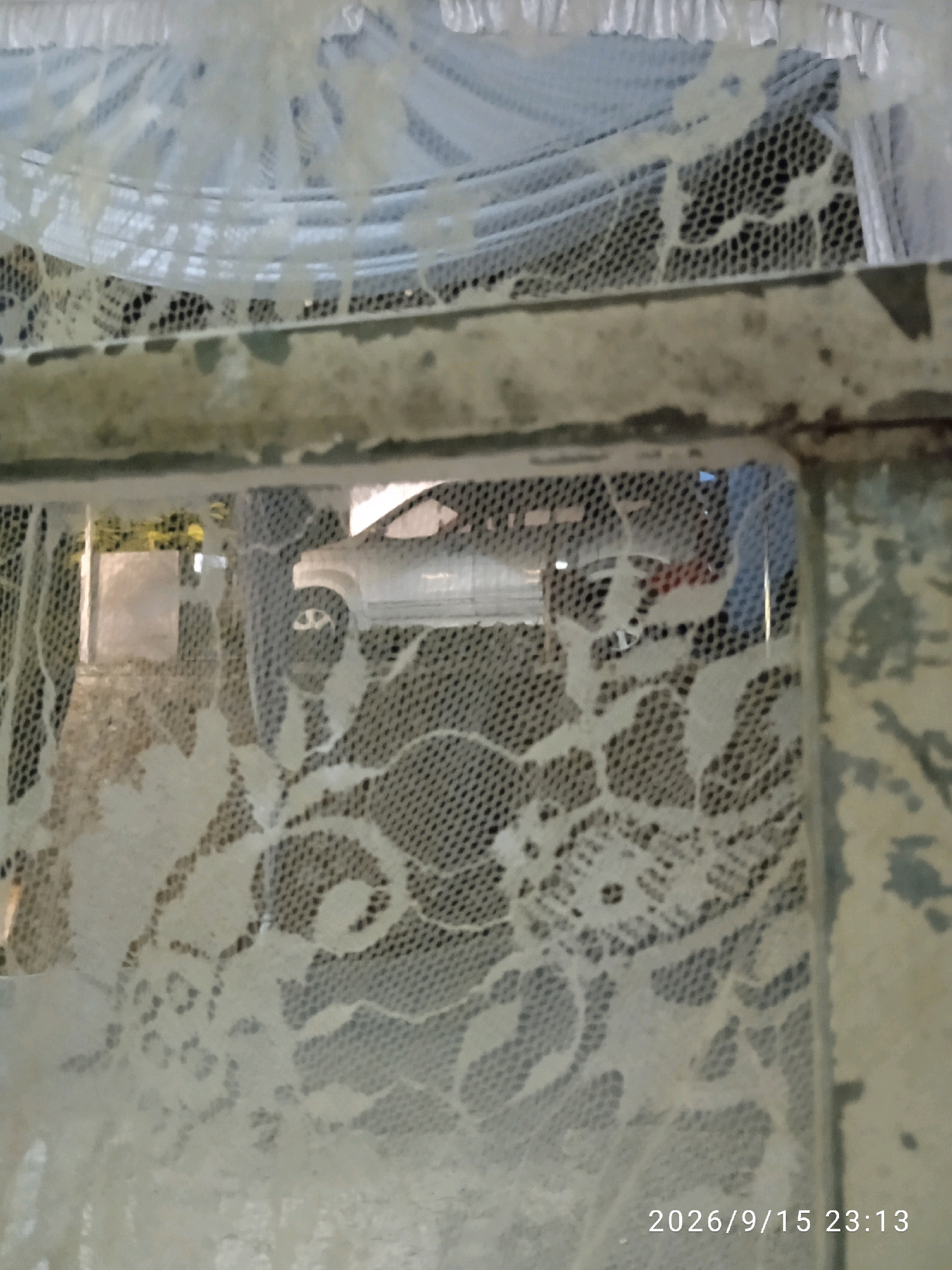 A close-up shot shows a white lace curtain draped over a window frame. The lace is intricate, with floral patterns. Through the lace, a portion of a white car is visible, suggesting it is parked outside or passing by. The background is blurred and indistinct, hinting at an urban environment. A timestamp, "2026/9/15 23:13", is visible in the lower right corner of the image, indicating the date and time the photo was taken. The location is not specified in the image.