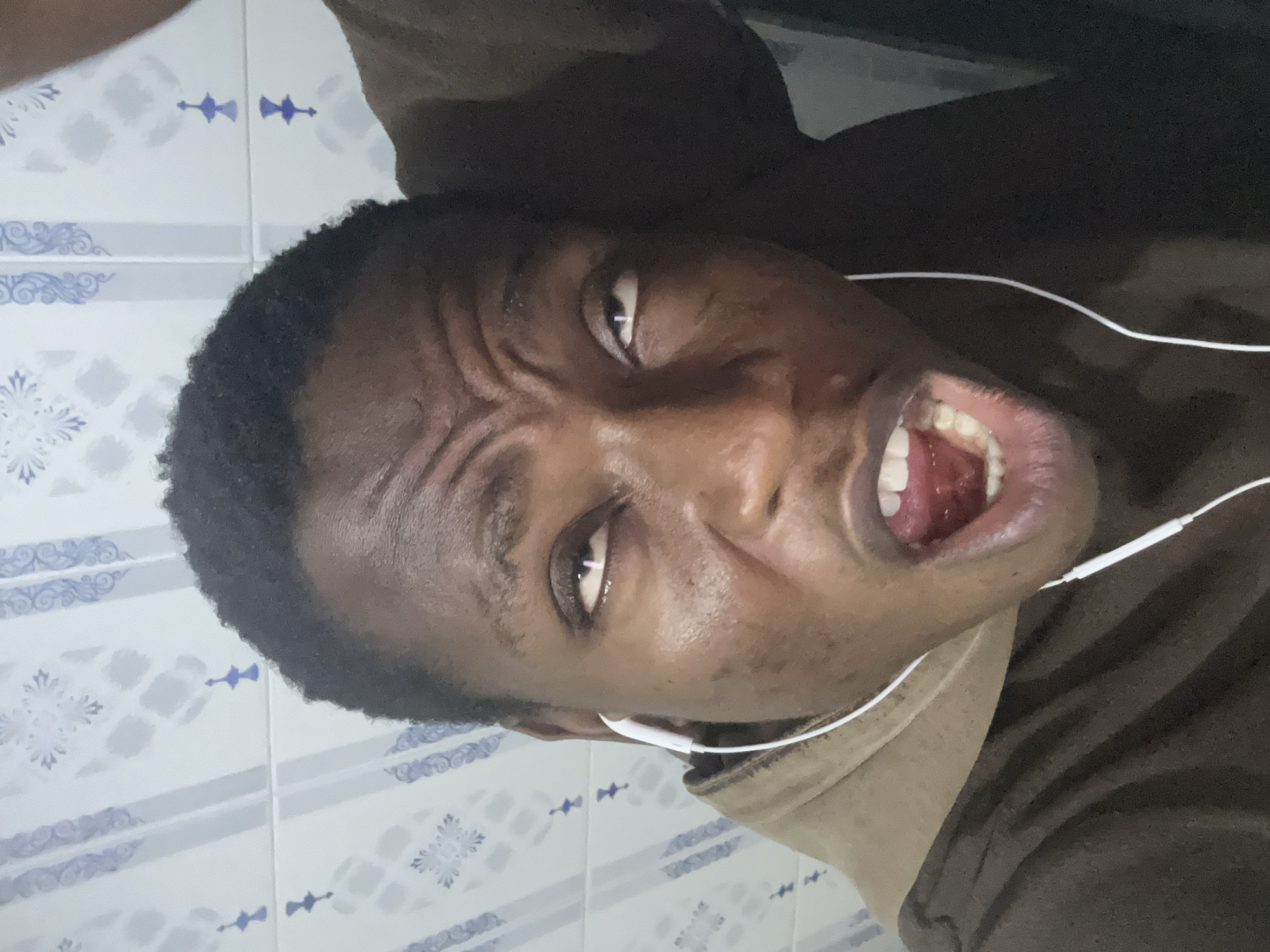 A close-up, slightly distorted image captures a dark-skinned individual with short, dark hair, wearing white earbuds. Their mouth is open wide, revealing teeth and the interior of their mouth. The person's facial expression is intense, with furrowed brows. The background appears to be a tiled wall, possibly in a bathroom, with a repeating blue and white floral-like pattern. The location is Unknown, Unknown.
