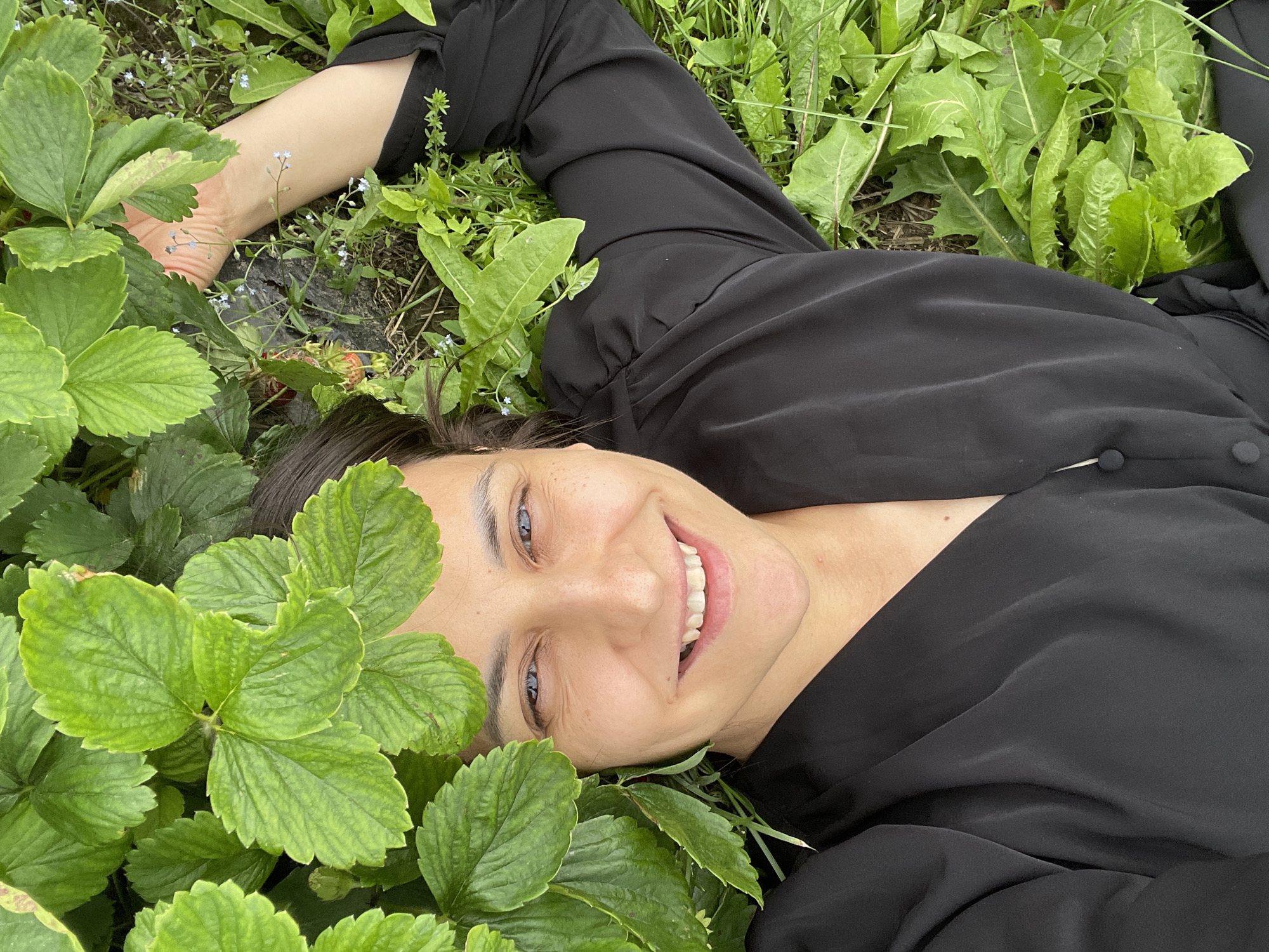 The media file features a close-up, overhead shot of a woman lying on her back amidst lush greenery, likely in a field or garden. She is wearing a flowing black top. Her head is tilted slightly, and she is looking up at the camera with a broad, genuine smile, her eyes wide and bright blue. Some of her dark hair is visible peeking out from under the foliage. Green strawberry leaves frame her face, and other wild green plants and some small blue wildflowers are interspersed around her. Her left arm is bent, with her hand resting on the ground among the plants, showing her bare arm and wrist. The lighting suggests it is daytime, possibly overcast or diffused sunlight, creating a soft glow on her face. The setting appears natural and outdoors, possibly in Niva, Russia, given the context. The overall emotion conveyed is one of joy, contentment, and connection with nature. No text is visible in the media file.