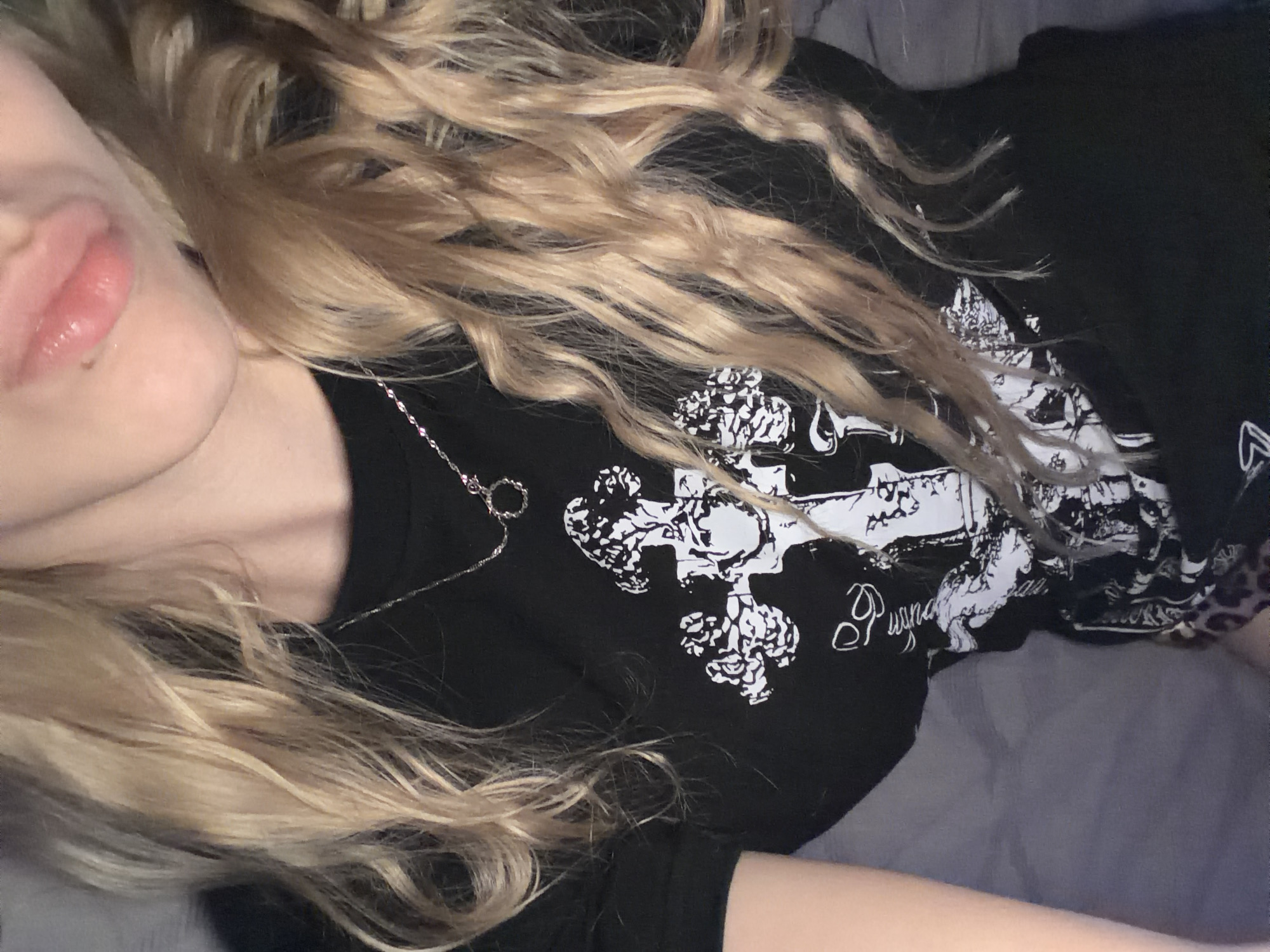 A close-up, overhead shot shows a young woman with long, blonde, wavy hair. She is wearing a black t-shirt with a white graphic design featuring a cross. A thin silver necklace with a circular pendant is visible around her neck. Her lips are slightly parted and appear to have a glossy finish. The background consists of a grey surface, likely a bed or couch. The image is cropped to focus on the upper torso and face of the subject.