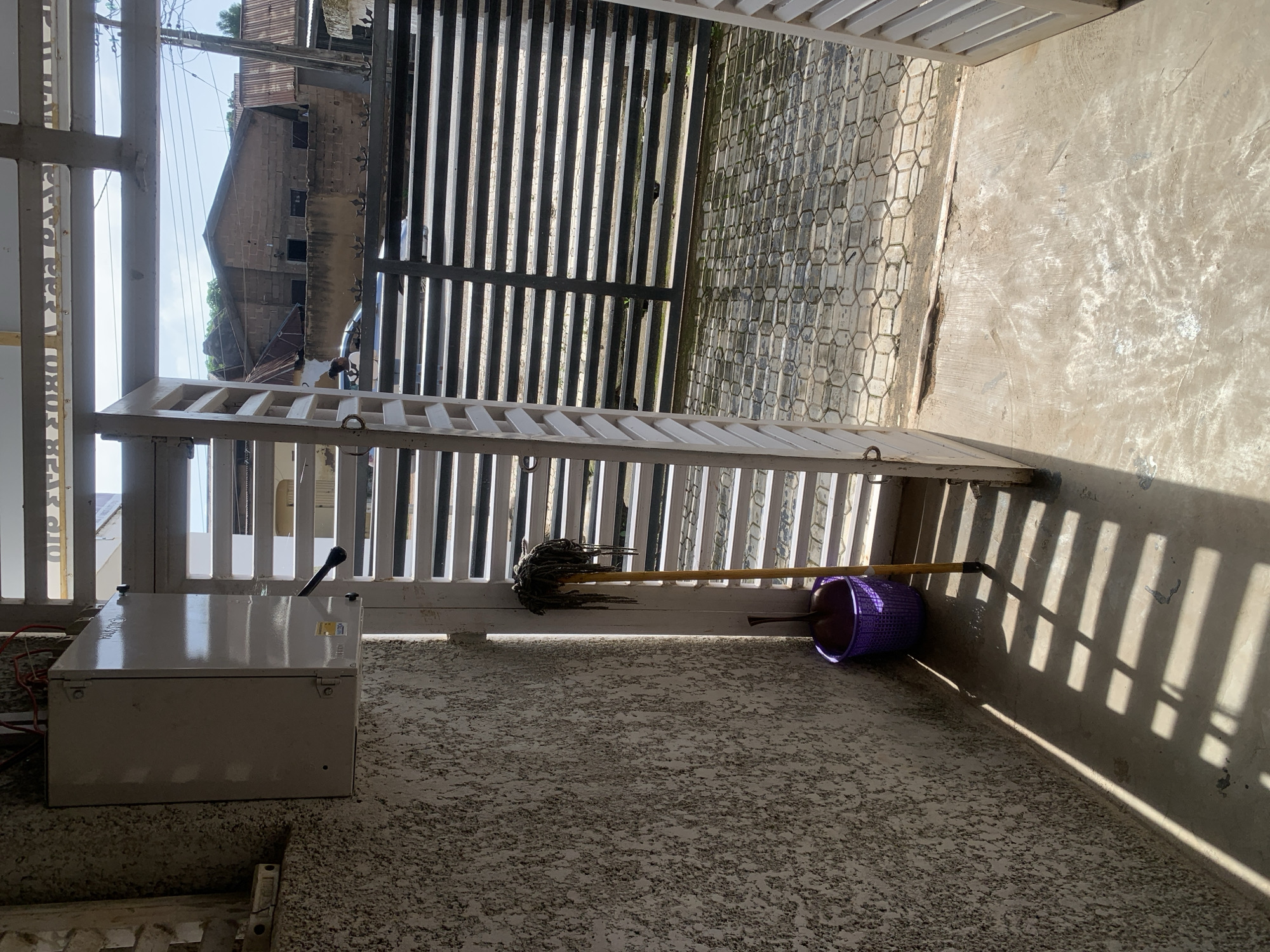 An outdoor view looking out from an entryway onto a paved area with buildings in the background. On the left, a textured wall has a light-colored electrical box mounted on it. Next to the wall, a yellow mop with a dark mop head stands upright in a purple woven basket. Sunlight casts striped shadows on the concrete floor. The entryway is framed by white slatted doors which are open, revealing a metal slatted gate beyond. The paved area outside is made of interlocking hexagonal stones. In the background, brick buildings with corrugated metal roofs are visible under a clear blue sky.