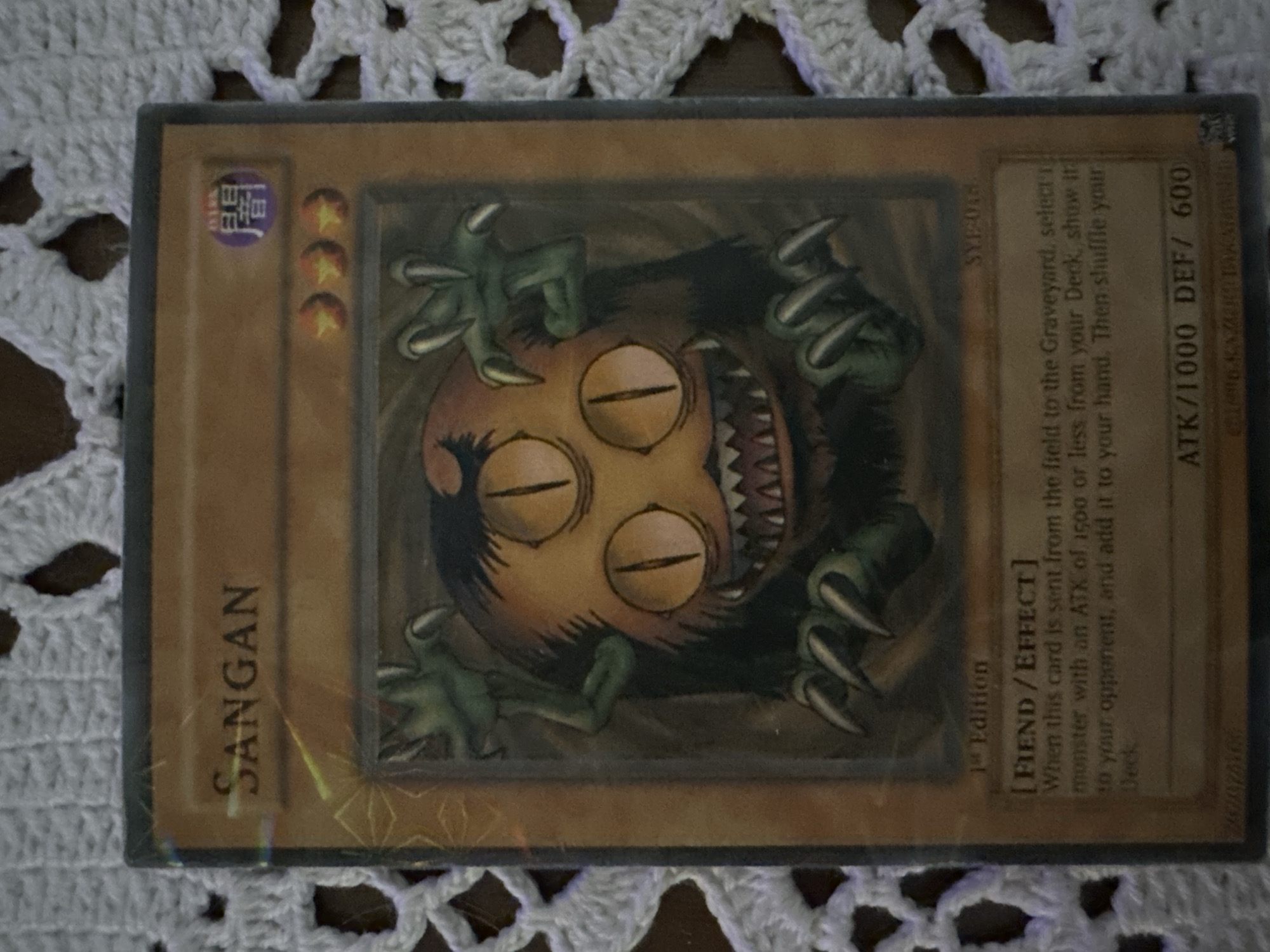 A Yu-Gi-Oh! trading card for the monster "Sangan" rests on a white crocheted surface. The card is oriented diagonally with the title of the card written in English with Japanese characters at the top left. The art is dark with the monster appearing to reach out from the center. A 1st edition stamp is present on the card as well as its text. The card's attack and defense stats are located on the bottom right corner. The photo was taken in an indoor environment under bright lighting. There is no indication of the city of Sofia, Bulgaria.