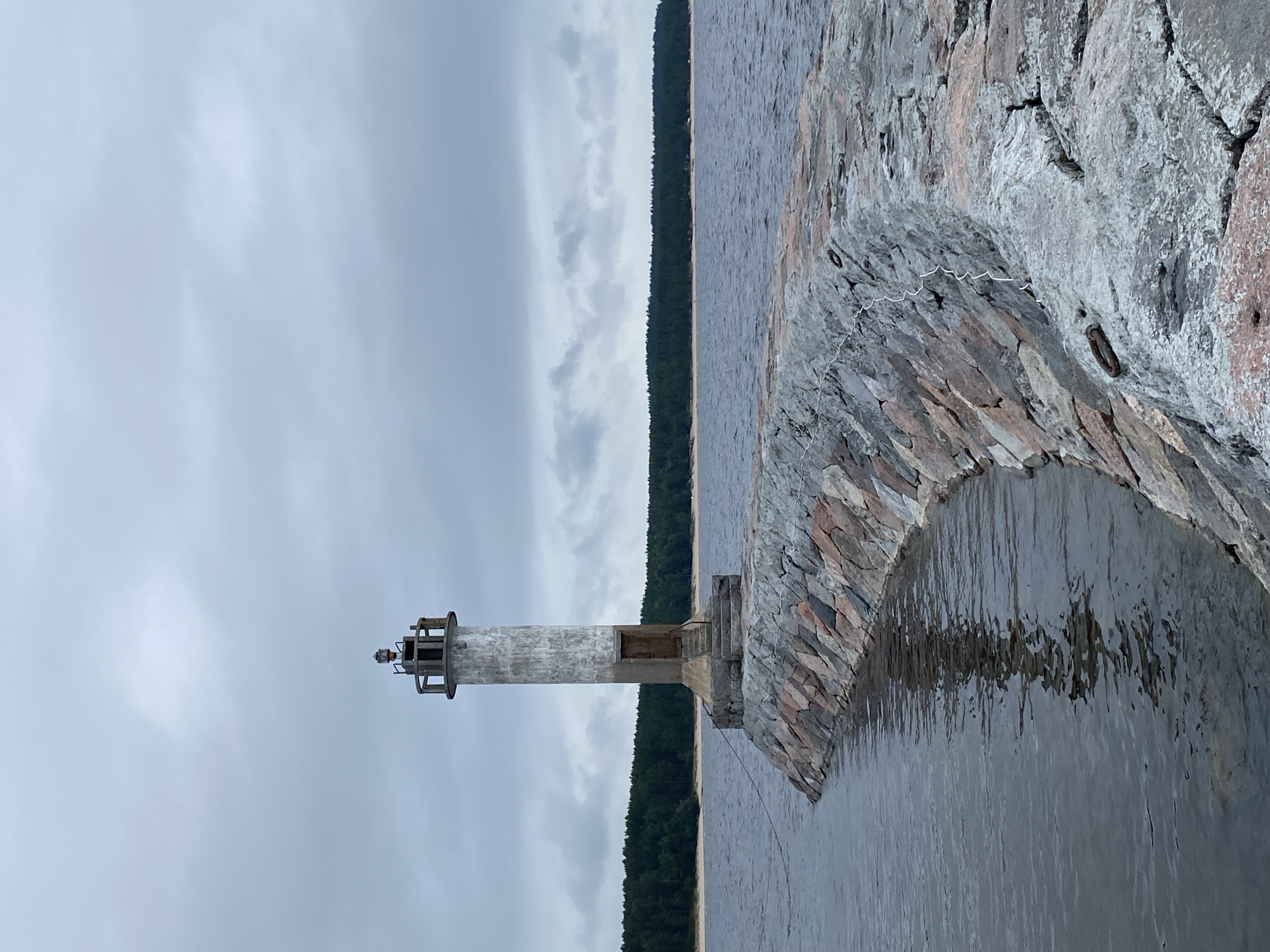 This is a photograph of a lighthouse situated on a stone pier extending into a body of water. The lighthouse is cylindrical and made of concrete, with a weathered appearance and a small entrance door at its base. It is topped with a lantern room. The pier is constructed from large, irregular stones of varying colors, including pinkish-red and gray. The water is dark and somewhat choppy, reflecting the cloudy sky. In the background, a dense green forest lines the opposite shore, with a strip of sand visible along the waterline. The sky is overcast with gray clouds. The scene is located near the settlement of Моторное, Russia. There are no people visible in the image.