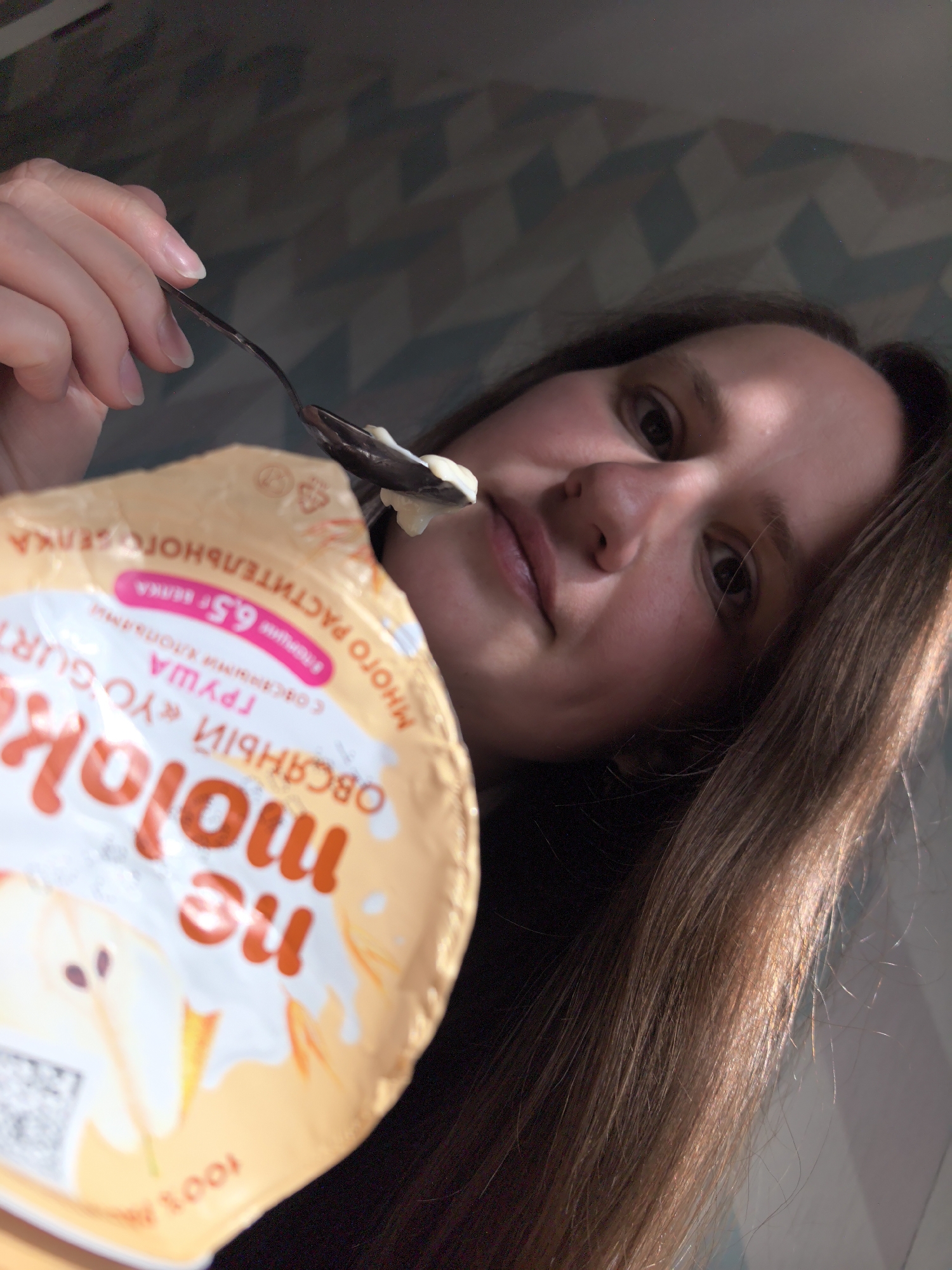 A woman is holding a spoon with yogurt in front of her face. She is positioned to the right of the yogurt cup. The cup's label reads "ne molok," which translates to "not milk." The label also indicates that it is an oat-based yogurt with pear flavor. The text on the label is in Russian.
The location appears to be indoors, as a wall with a geometric pattern is visible in the background. It's not possible to confirm the exact location in Фролы, Russia, from the image alone.