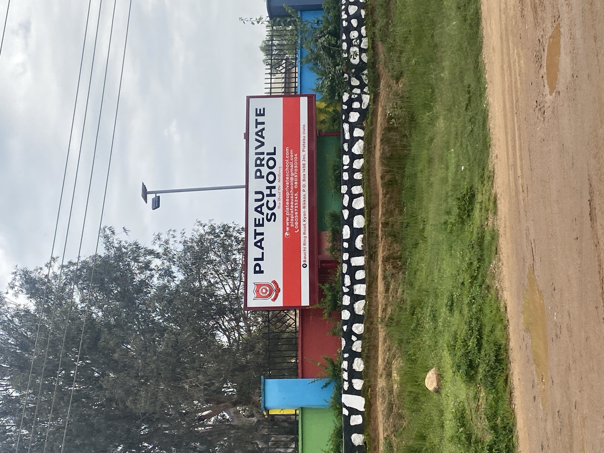 A weathered sign for "PLATEAU PRIVATE SCHOOL" stands prominently in front of a school building. The sign is predominantly white with red accents and features the school's emblem, website address, email, and phone numbers. The school appears to be located in Jos, Nigeria, as indicated by the text on the sign which reads "Bauchi Ring Road, Kyan-Rikkos, P.O. Box 1498 Jos, Plateau state." The entrance to the school is bordered by a colorful wall in shades of green, blue, and red, and a black metal fence. A decorative black and white pattern is visible along the base of the wall. Behind the school, trees and utility wires are visible against a cloudy sky. In the foreground, a dirt road leads towards the school, with patches of grass and a small puddle of water on the road. A single rock sits in the grassy area to the left.