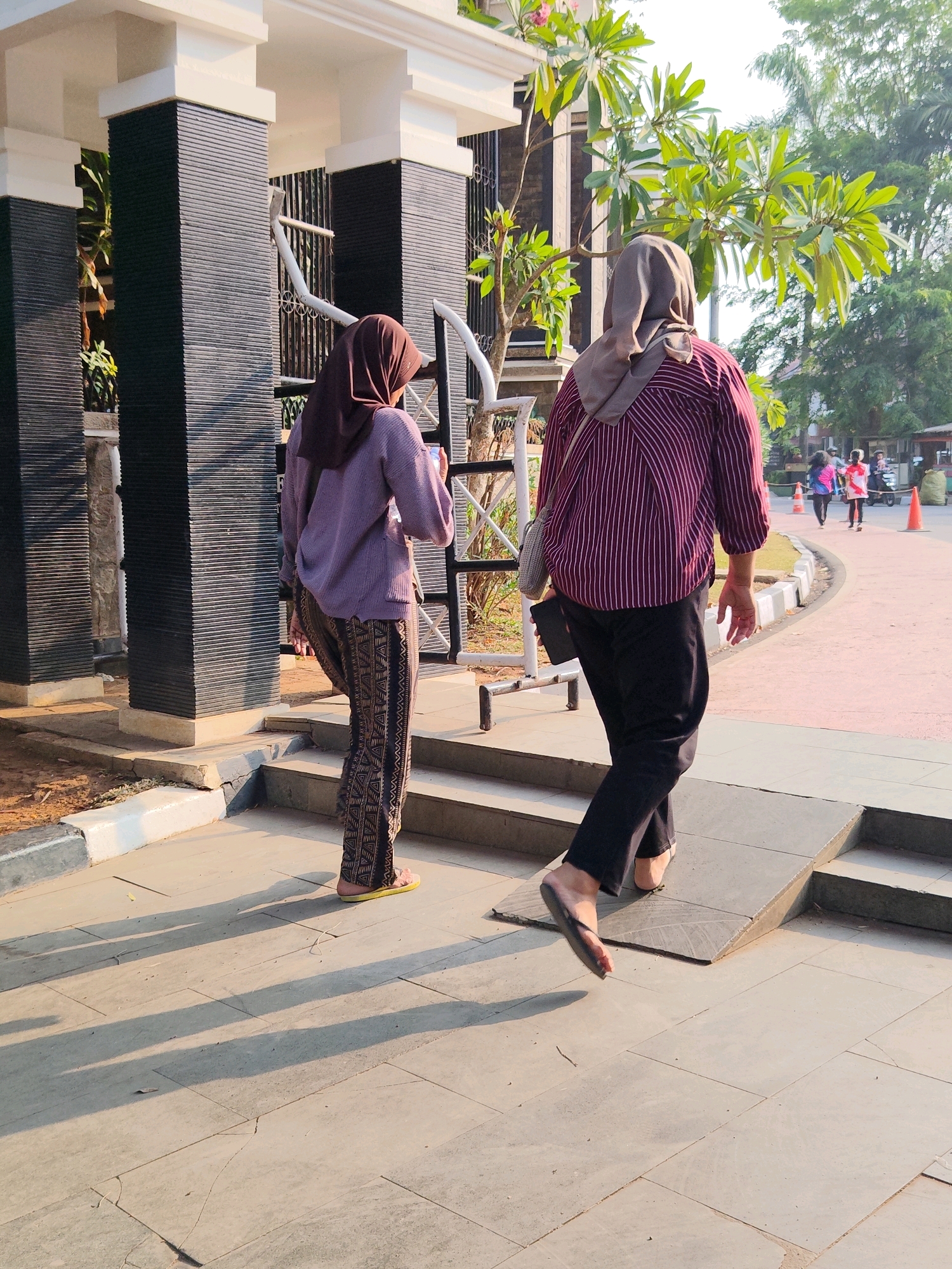 Two women, facing away from the camera, walk on a tiled pathway in Purwakarta, Indonesia. The woman on the left wears a purple sweater and patterned pants. The woman on the right wears a striped shirt, black pants, and a grey hijab. A ramp is visible in the foreground. In the background, other people walk on a sidewalk lined with traffic cones. Trees and buildings are also visible. The scene is illuminated by bright sunlight, casting long shadows on the ground.