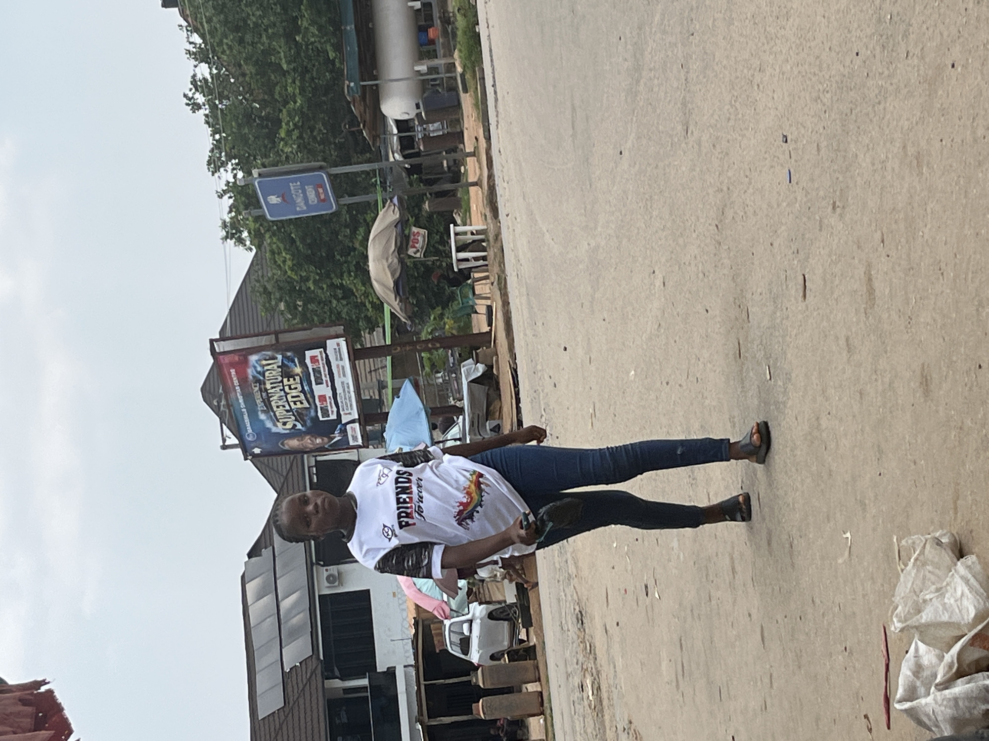 This media file shows a young woman standing on a dirt road. She is wearing a white t-shirt with "FRIENDS forever" printed on it in black and red, along with a colorful rainbow graphic. She also wears dark blue jeans and dark sandals. She is looking towards the camera with a neutral expression.
The scene appears to be a street in Abuja, Nigeria, with buildings and trees visible in the background. A large billboard with the words "SUPERNATURAL EDGE EXPERIENCE" is prominently displayed behind the woman. There are also signs that read "DANGOTE CEMENT" and "POS". The ground is dusty and uneven, with some debris scattered around, including a white plastic bag. A white vehicle is parked nearby. The overall lighting suggests it is daytime, likely overcast given the soft light. The environment appears to be a bustling commercial or residential area.