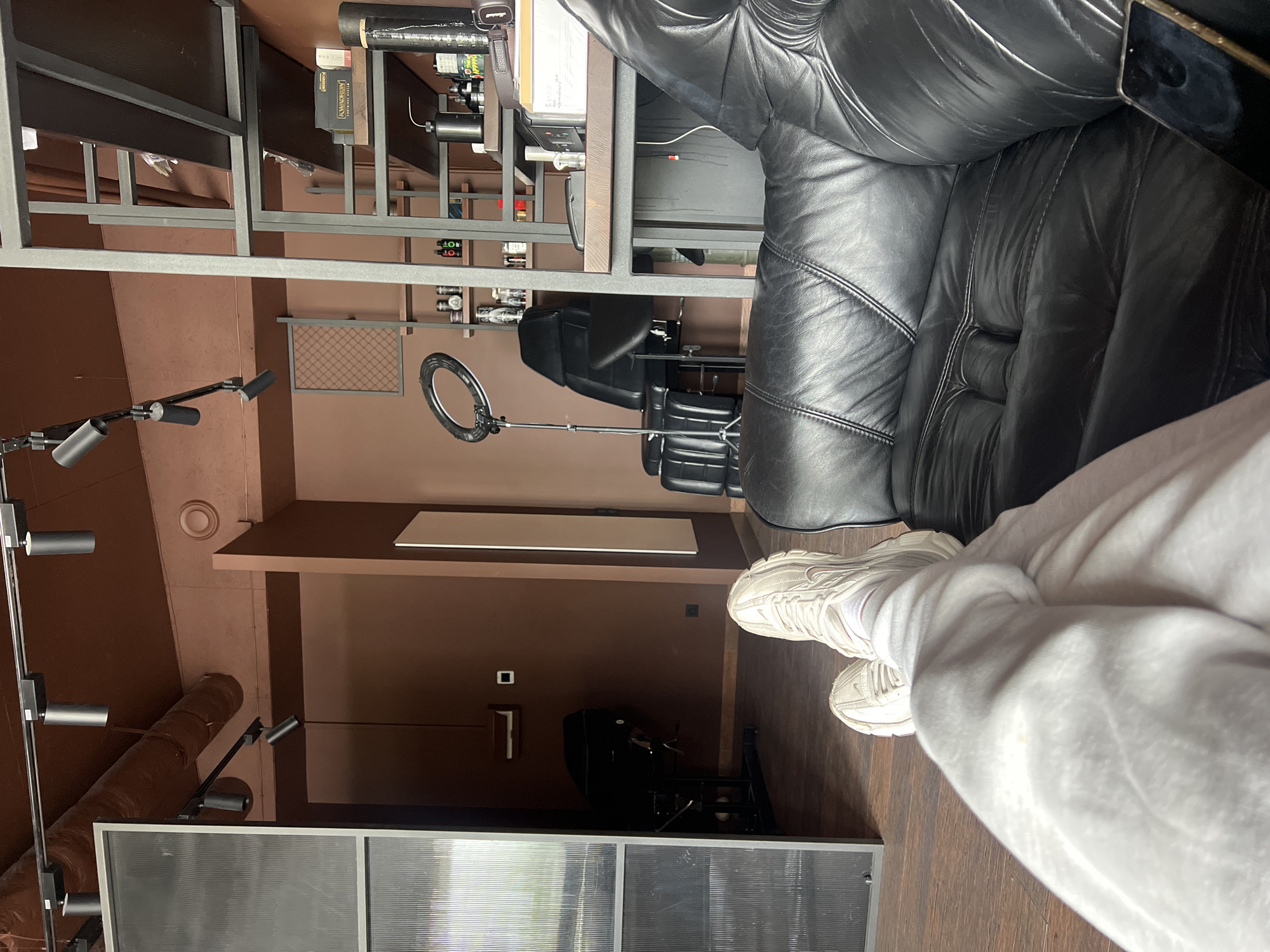 The scene is an indoor space with a modern, slightly industrial aesthetic. The primary subject is a person wearing light gray sweatpants and white sneakers sitting on a black leather sofa. The sofa is plush with quilted detailing. The background includes a multi-tiered metal shelf unit containing various items. Further into the background, there is a salon-style setup with a black reclining chair and a ring light. The walls are a muted brown color. In the lower left corner, there is a partially visible glass partition. The location is not explicitly specified as Ternopil, Ukraine.
