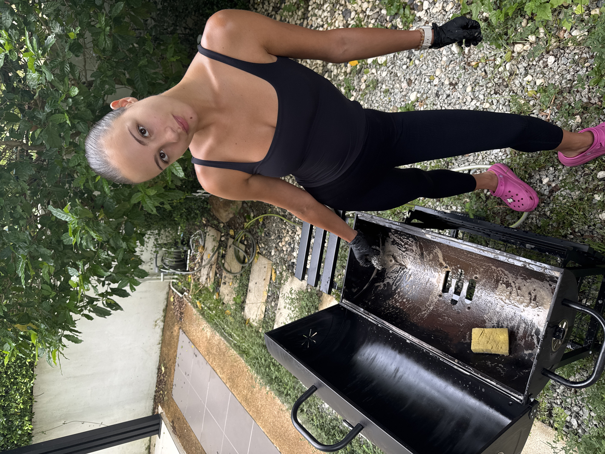 A young woman with dark hair pulled back, wearing a black tank top, black leggings, and pink Crocs, is cleaning a black charcoal grill in an outdoor garden setting in Chalong, Thailand. She is wearing black gloves and is using a yellow sponge and a scrubbing brush to clean the inside of the grill, which appears to have soapy water and residue. The grill is open, revealing its interior. The background shows lush green foliage, a white wall, a tiled pathway, gravel, and a coiled hose. The scene suggests a domestic chore being performed in a residential area.