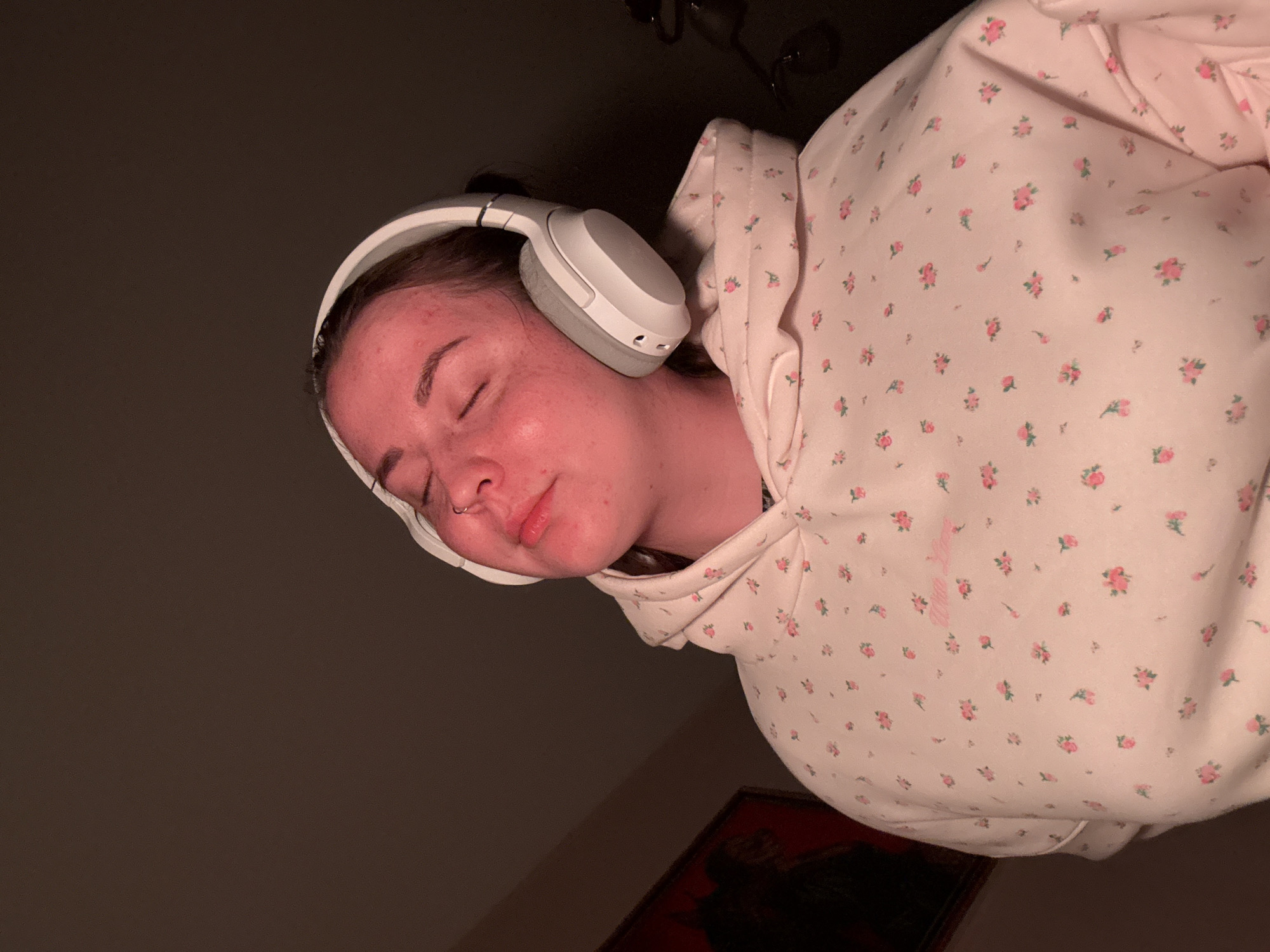A person is wearing headphones and a floral hoodie. They are lying down with their eyes closed. The scene is indoors, and the background is dark. The location is not specified, so I cannot comment on whether it is in Izhevsk, Russia.