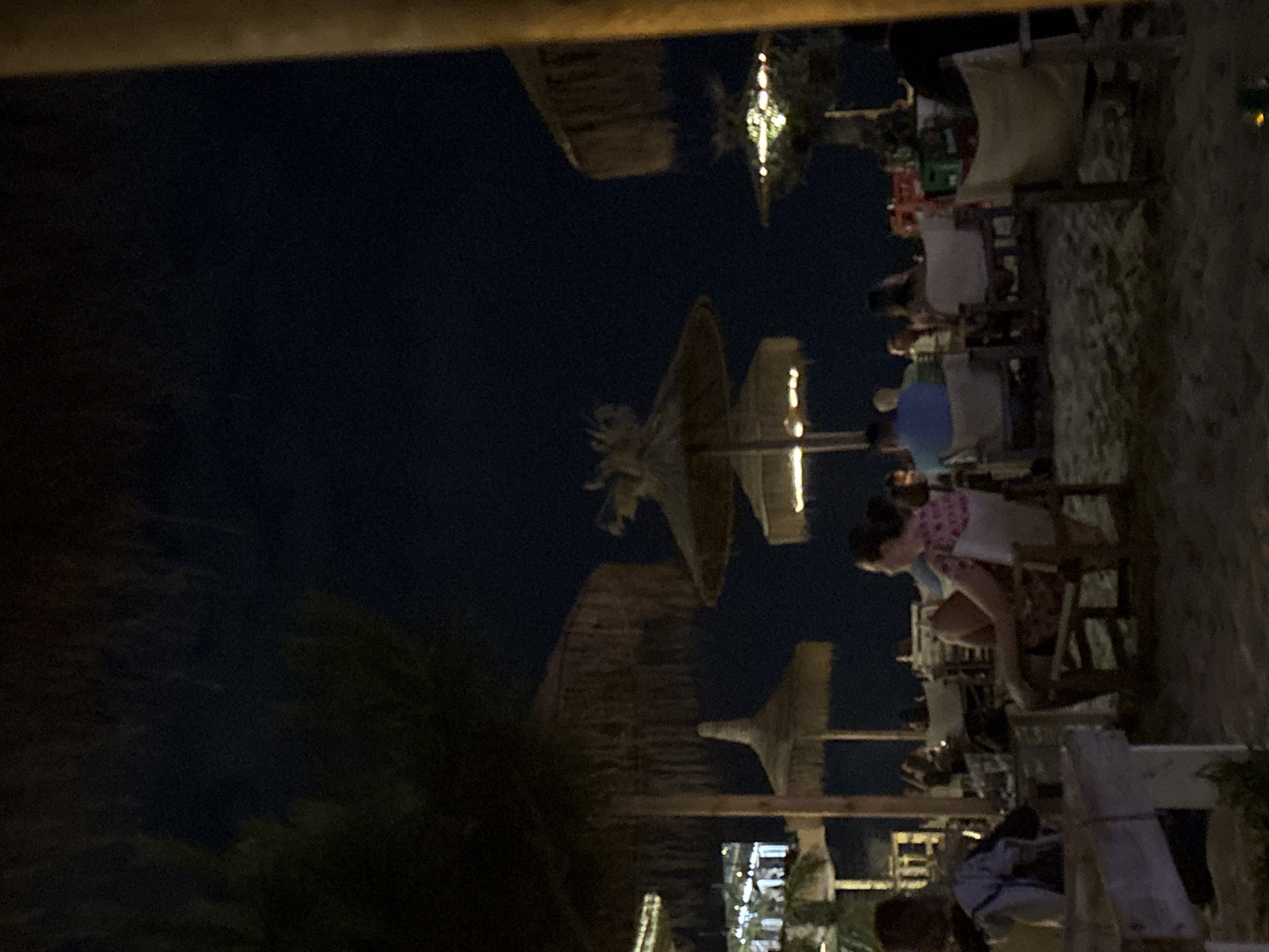 The image depicts an outdoor seating area at night, likely a beachside restaurant or bar. The ground is sandy, and several thatched umbrellas provide shade. In the foreground, a wooden structure, possibly a table or bench, is visible. Further back, people are seated at tables and in chairs, some interacting with each other, others looking outwards. The lighting is dim, suggesting artificial illumination from the establishment, with some subtle reflections on surfaces. The sky is dark, indicating nighttime. The overall atmosphere is relaxed and social.