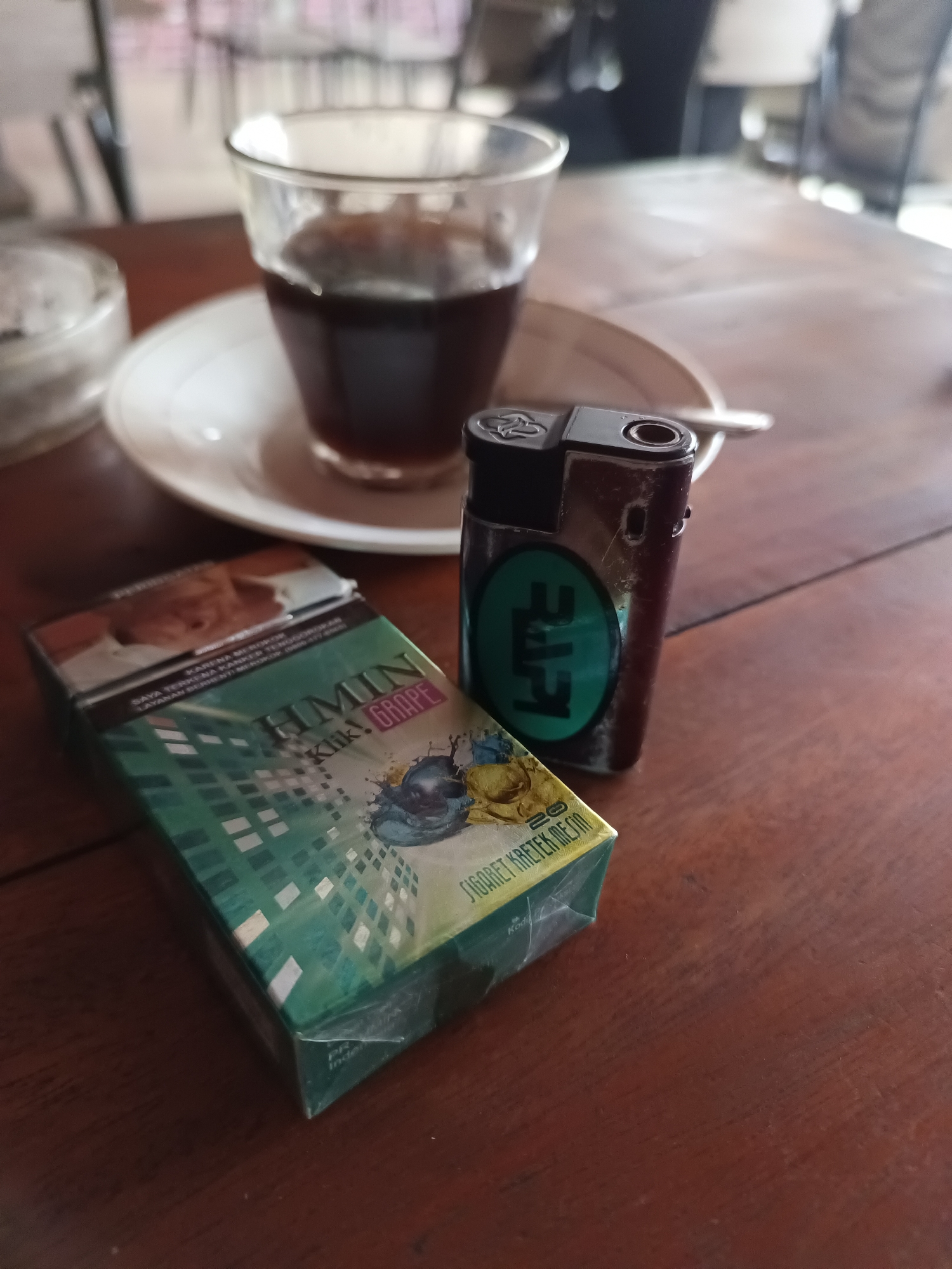 This is a medium-angle shot of a wooden table with a pack of cigarettes, a lighter, and a glass of dark liquid, likely coffee or tea, on a saucer. The cigarettes are from a pack labeled "HMIN Klik GRAPE" and feature green packaging with a graphic of a beetle. The lighter is dark-colored with a green circular logo containing the letters "LTE." The glass of beverage is partially filled. In the background, the scene is blurred, suggesting an indoor setting like a cafe or home. The overall mood is relaxed and casual, possibly during a break or a moment of quiet contemplation. The visible text on the cigarette pack includes warnings about smoking and indicates that it is a machine-rolled kretek cigarette. The location is contextually identified as Lhokseumawe, Indonesia.