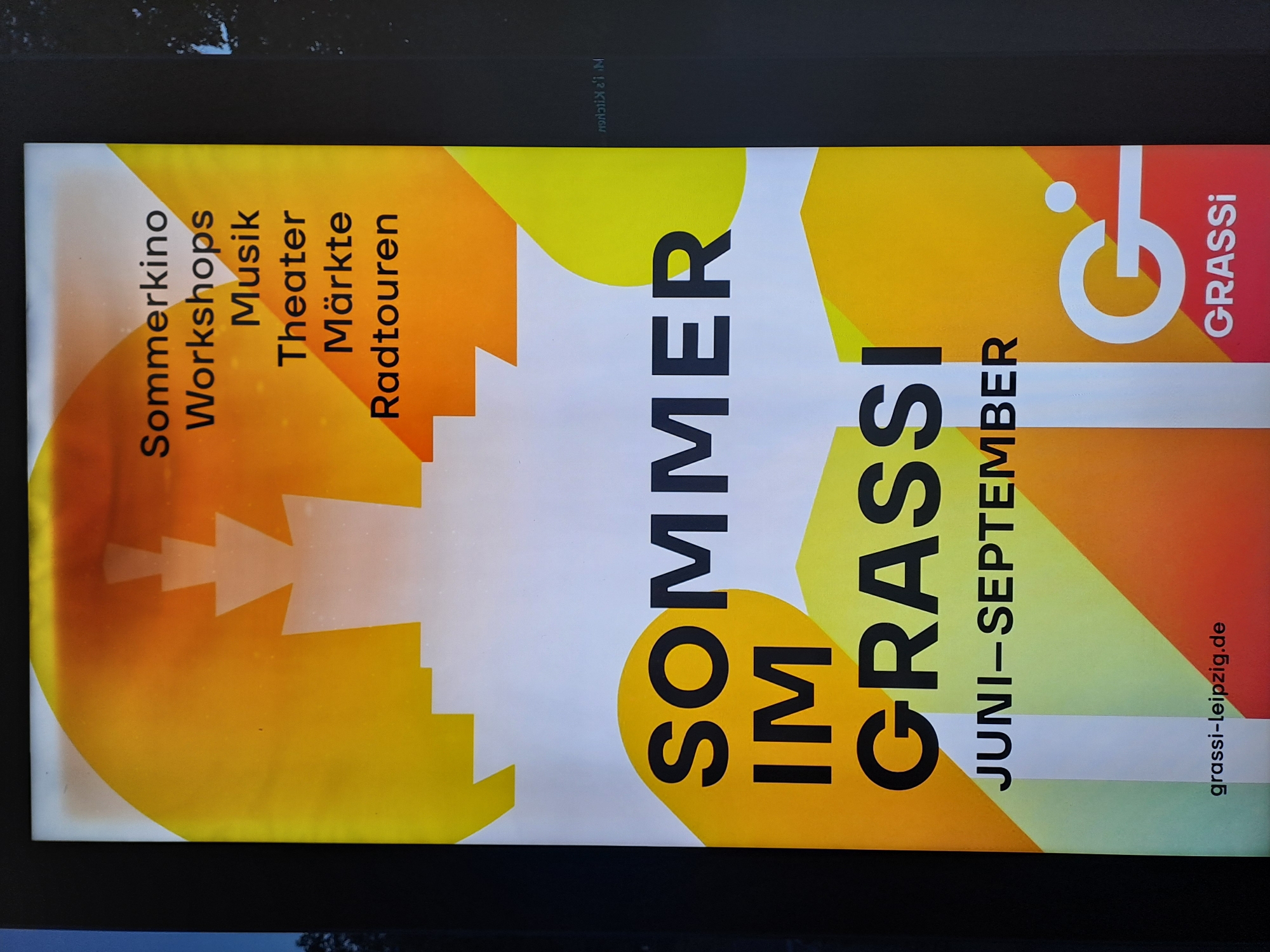 A poster for "SOMMER IM GRASSI" (Summer at Grassi) is displayed, featuring a bold orange and yellow abstract design with white shapes resembling buildings or a cityscape. The text lists various events: "Sommerkino," "Workshops," "Musik," "Theater," "Märkte," and "Radtouren." Below this, the central title reads "SOMMER IM GRASSI" in large black letters, followed by the dates "JUNI-SEPTEMBER." At the bottom, a website address "grassi-leipzig.de" and a logo with "GRASSI" are visible. The poster is positioned outdoors, likely in a public space in Leipzig, Germany, as indicated by the website address which refers to the Grassi Museum für Angewandte Kunst in Leipzig. The poster appears to be illuminated by natural light.