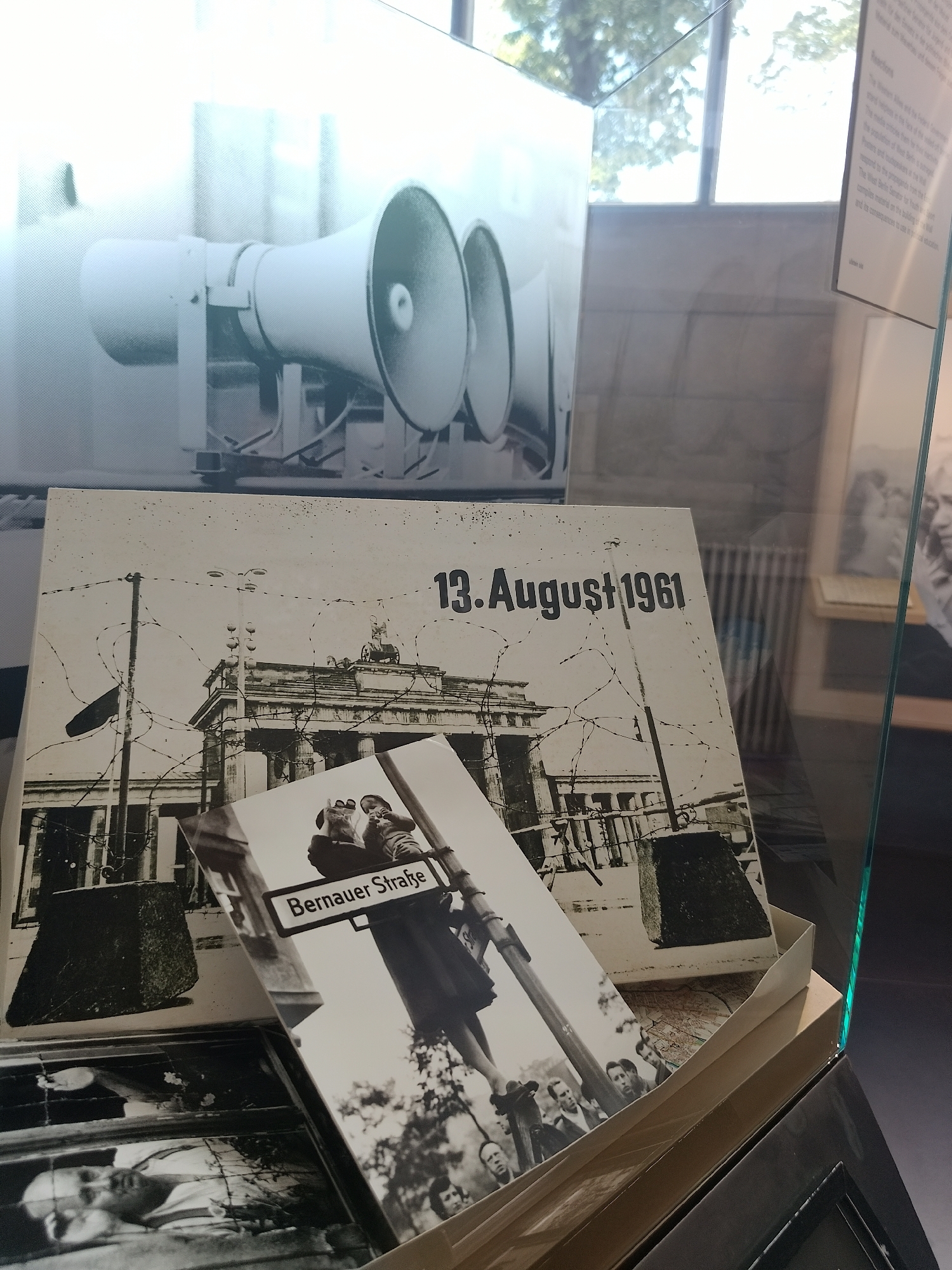 A display inside a museum exhibit showcases photos related to the Berlin Wall. In the background, a black and white photo of loudspeakers is displayed. The foreground contains a box with images, including one of the Brandenburg Gate with barbed wire and the date "13. August 1961." A smaller photo on top reads "Bernauer Straße" and depicts people looking up, potentially at someone escaping. Additional photos are visible inside the box, featuring individuals. The exhibit is enclosed in glass, with reflections of the outside environment visible.