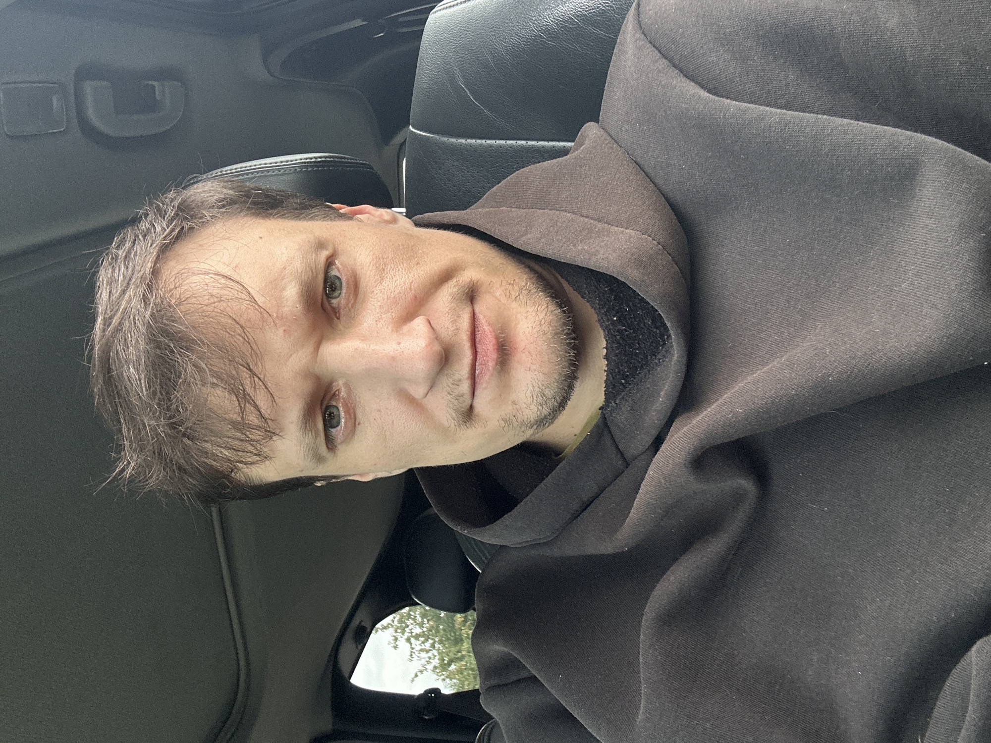 A man is captured in a close-up selfie taken from inside a car. He is seated in the driver's seat, facing the camera directly. The man has light skin, light brown hair, and light blue eyes. He is wearing a dark brown hoodie with the hood up. The interior of the car is visible, including the car's ceiling with a sunroof, and the leather headrest of the passenger seat. Outside the car window, trees and foliage are visible, suggesting a natural or suburban setting. The photograph was taken in Makiv, Ukraine.
