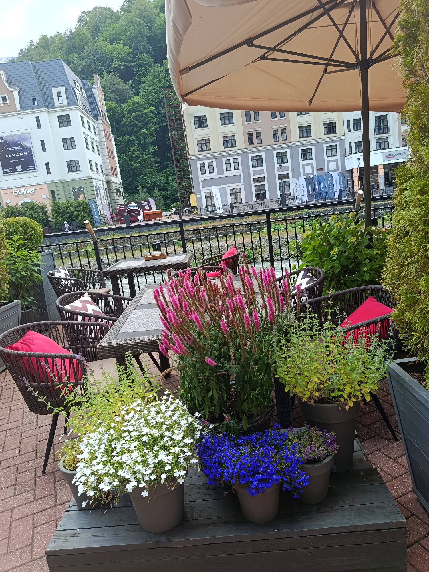 This media file depicts an outdoor seating area for a cafe or restaurant in Estosadok, Russia, overlooking a river. The scene is set under a large, cream-colored umbrella, providing shade. Several woven chairs with red and patterned cushions surround wicker tables. In the foreground, there are pots of flowers, including vibrant pink spikes, delicate white blooms, and bright blue clusters.
Behind the seating area, a black metal fence separates the patio from a river. In the background, there are multi-story buildings with facades in shades of white, beige, and gray, featuring numerous windows and balconies. The architecture suggests a resort town. Lush green trees and a hillside rise behind the buildings, indicating a mountainous or forested environment.
There are no people prominently featured in the image, though a few figures can be seen walking in the distance near the buildings. The weather appears overcast, suggesting a cool or temperate day. The overall mood is peaceful and inviting, emphasizing a relaxing outdoor dining experience.
Visible text includes "Gulliver" on the buildings in the background. Additional text appears on banners further in the background, but it is illegible at this resolution. The image captures a serene moment, highlighting the interplay of nature and architecture in a scenic location.