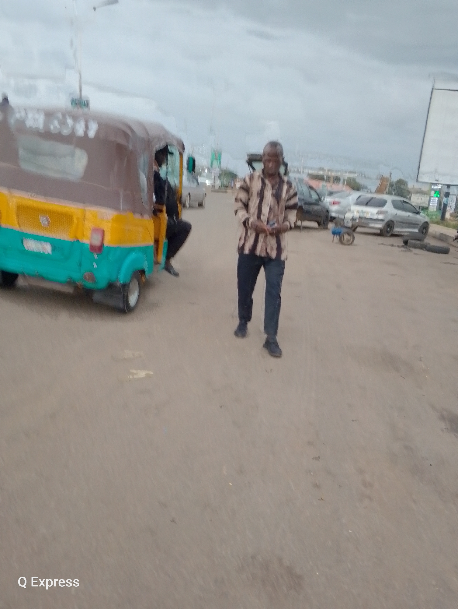The image depicts a street scene in Jos, Nigeria, under an overcast sky, suggesting it is either early morning or late afternoon, or possibly a cloudy day. The foreground is dominated by a portion of a yellow and turquoise auto-rickshaw, a common mode of transport in many parts of Nigeria. The rickshaw appears to be stationary or moving slowly.
To the right of the auto-rickshaw, a man is walking on the dusty road. He is wearing a striped collared shirt and dark trousers, and appears to be holding something in his hands. His expression is not clearly discernible, but his posture suggests he is walking purposefully. Behind him, several other vehicles are visible, including a silver car and a darker vehicle. A small, wheeled contraption, possibly a generator or pump, is situated on the road near the silver car. In the background, there are buildings and vegetation, indicating an urban or semi-urban environment.
The overall scene suggests a typical day in a Nigerian city, with elements of daily life and commerce. The presence of the auto-rickshaw and the various vehicles on the road point to a functional street. The cloudy weather contributes to a muted color palette. At the bottom left corner of the image, the text "Q Express" is visible, which could be a watermark or branding related to the source of the image.