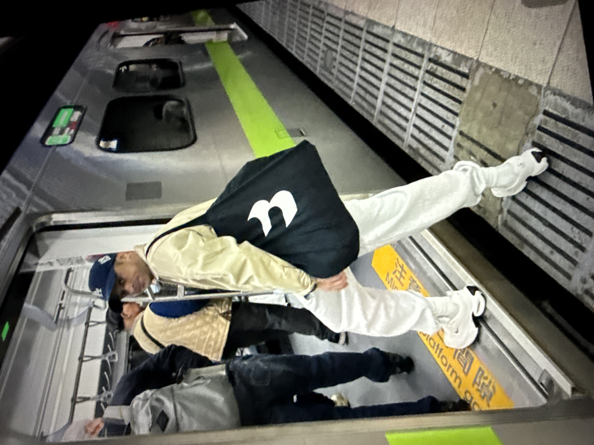 A person wearing a white tracksuit and white sneakers is standing on a train platform in Taipei, Taiwan. The person is holding a black bag with a white number '3' logo. They are facing away from the camera, looking towards the train. The train doors are open, and other passengers are visible inside. The platform is marked with yellow lines and has a metal grate. The train car is silver with a green stripe.