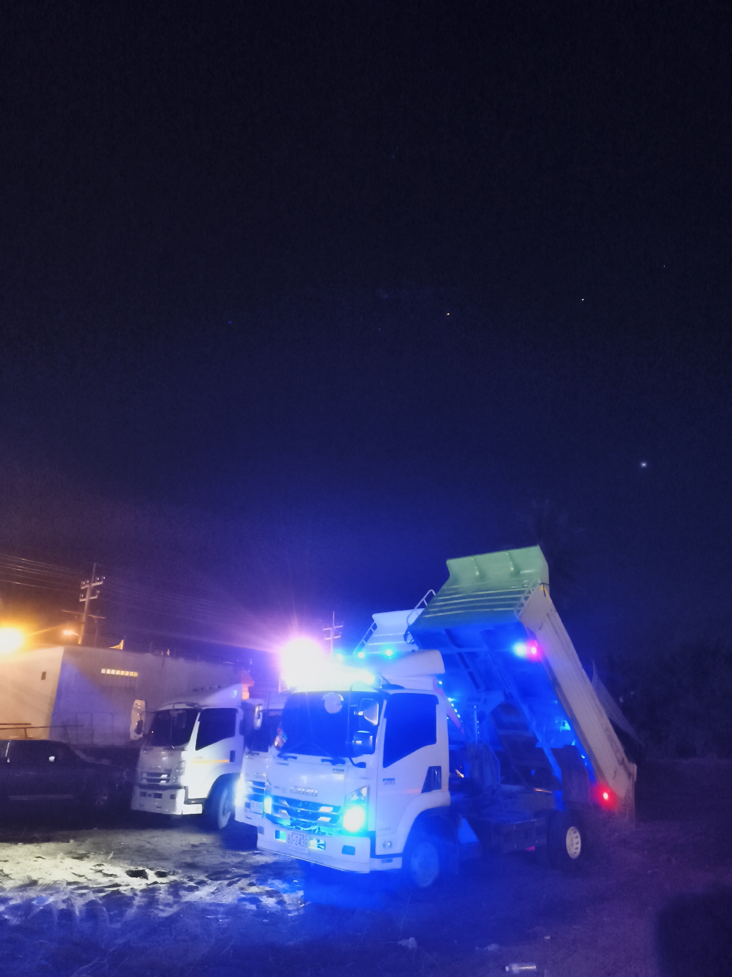 At night in Baan Mae Nam, Thailand, two white Isuzu dump trucks are parked with their dump beds raised. The closest truck is illuminated with blue lights. A third vehicle, possibly a car, is parked to the left near a building that's partially lit by a streetlamp. Overhead electrical wires are visible, and the night sky is dark with a few stars visible. The ground appears to be gravel or dirt.