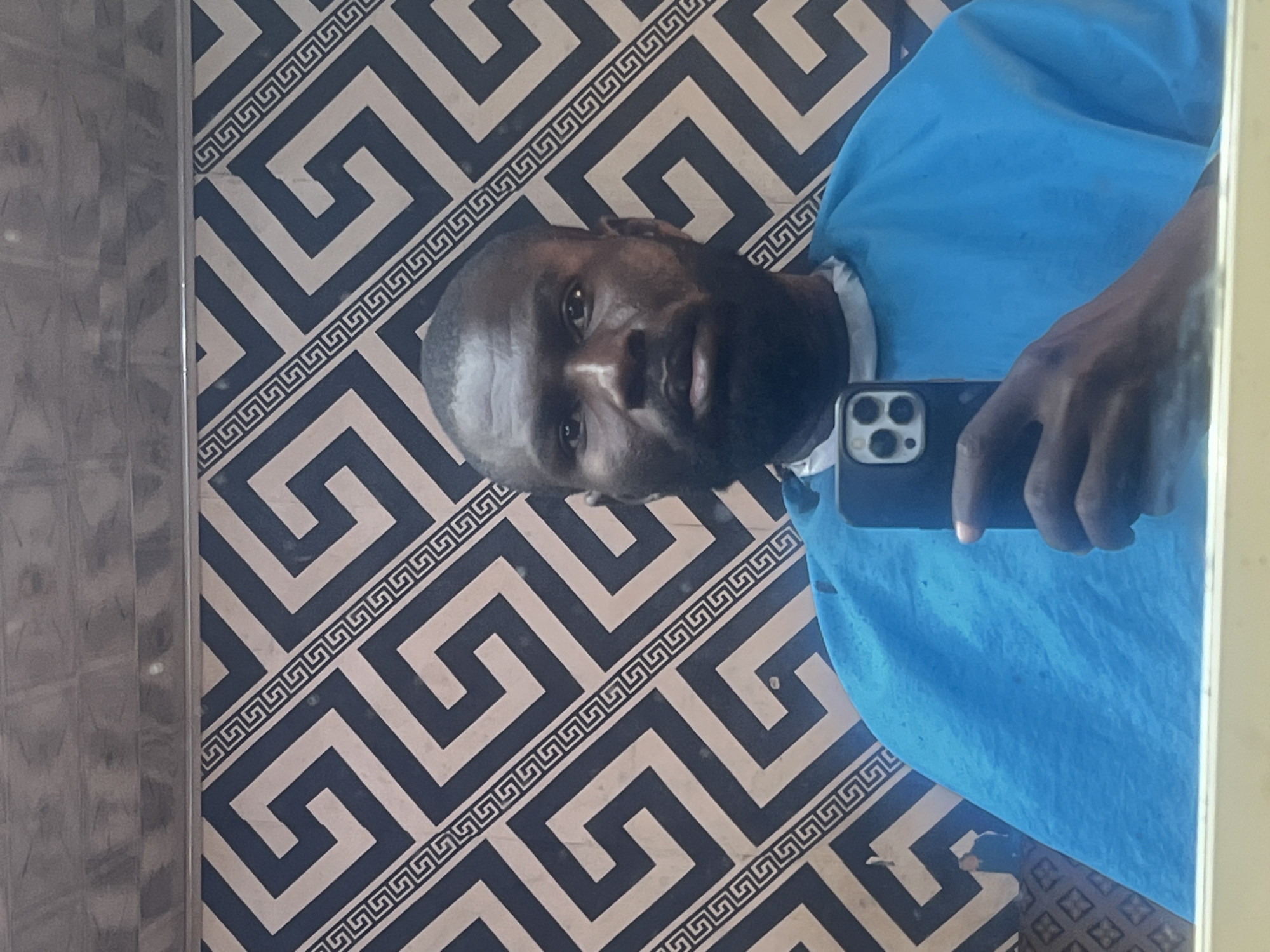 This image is a mirror selfie taken by a man, presented in a sideways orientation. The main subject is a man of African descent with short hair or a bald head and a beard, looking directly into the camera's reflection. He is wearing a blue short-sleeved top with a white collar band. He is holding a black smartphone, identifiable by its triple-camera module on the back, in his right hand, which is visible in the bottom right of the frame.
The background reflected in the mirror features a distinctive repeating geometric pattern resembling a Greek key or meander design, rendered in dark blue/black and light beige/tan colors, likely wallpaper or wall tiling. A narrow, vertically textured surface, possibly a column or wall feature, is partially visible on the left side of the frame. The overall setting appears to be indoors in Jos, Nigeria. The man's expression is neutral. There is no visible text in the image.