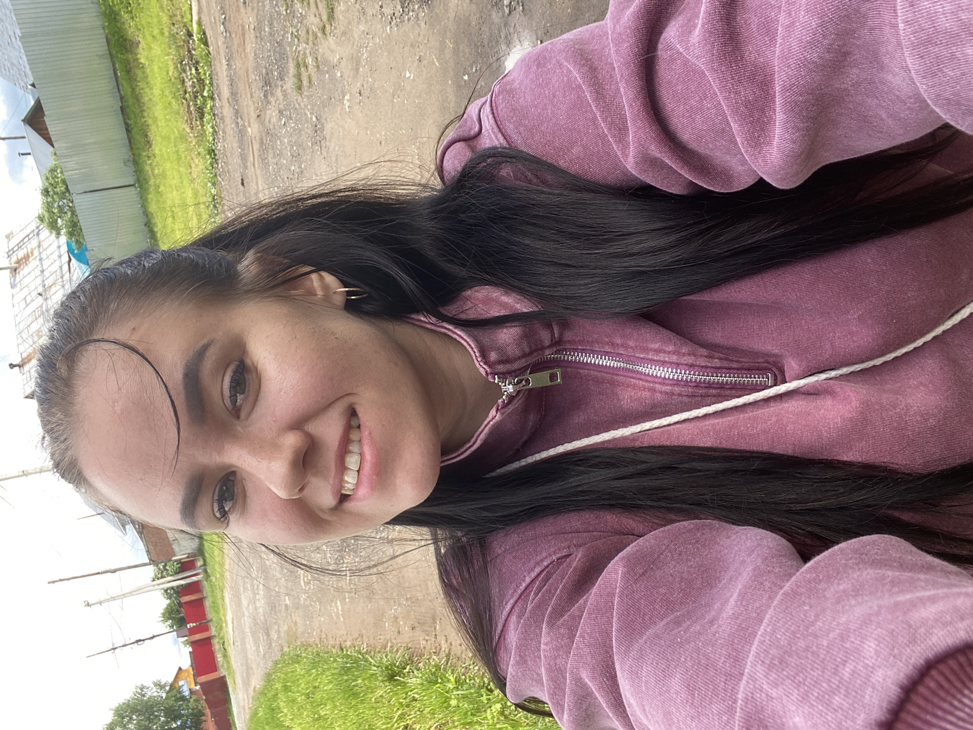A young woman is featured in a close-up, upward-angled selfie, smiling broadly at the camera. She appears to be in her late teens or early twenties, with long, dark brown or black hair, some strands falling across her forehead. She has dark, full eyebrows and visible eyelashes. Small, gold-colored hoop earrings are visible in both ears.
She is wearing a thick, textured hooded sweatshirt (hoodie) in a dusty rose or faded mauve color. The hoodie has a silver-colored zipper down the front and a white drawstring at the neckline.
The background suggests an outdoor, semi-rural setting, likely in Chaikovsky, Russia, as indicated by the location context. To the viewer's left, behind her right shoulder, is a dirt or gravel path/road. To the viewer's right, behind her left shoulder, is an area of bright green grass. Further back, there are various elements under a bright, possibly overcast daytime sky: a light-colored corrugated metal fence or building, several utility poles, a red fence, and some distant trees. The lighting is bright and even. There is no visible text in the image. The woman's expression is cheerful and pleasant.