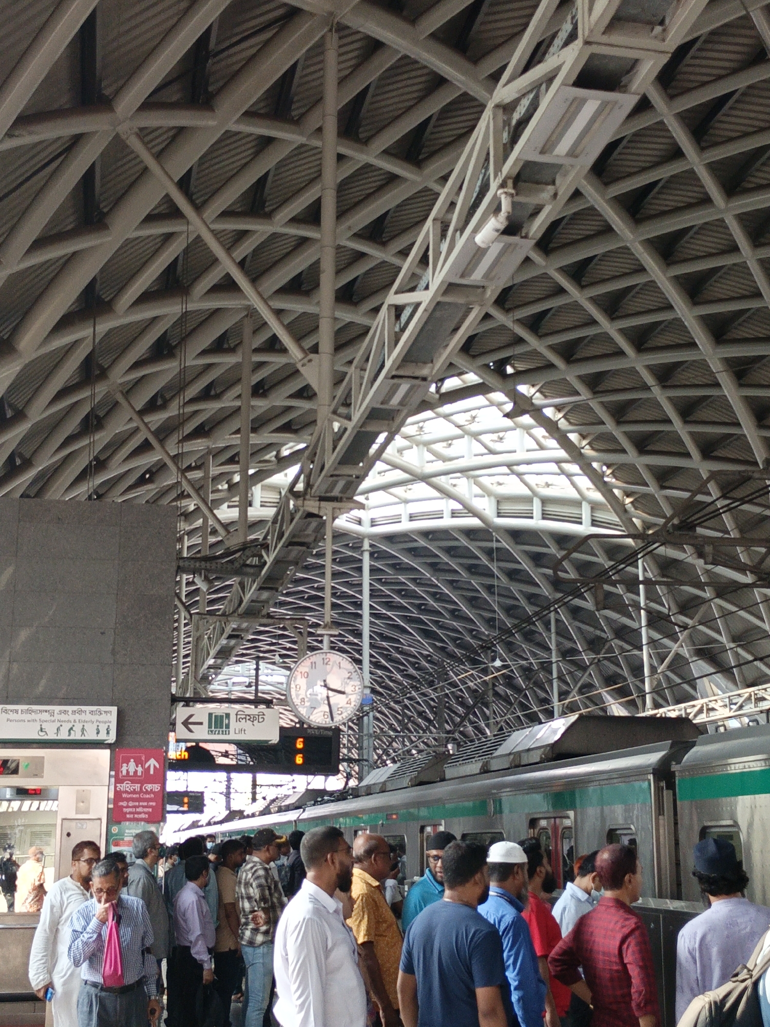 This indoor shot shows a crowded train station in Dhaka, Bangladesh. The architecture is dominated by a complex, crisscrossing metal structure forming the roof, allowing daylight to filter through. A large clock hangs from the structure. In the foreground, a group of people are waiting near a train. A sign with Bengali text and international symbols indicates facilities for special needs individuals and a women's coach. The scene suggests a bustling urban transportation hub.