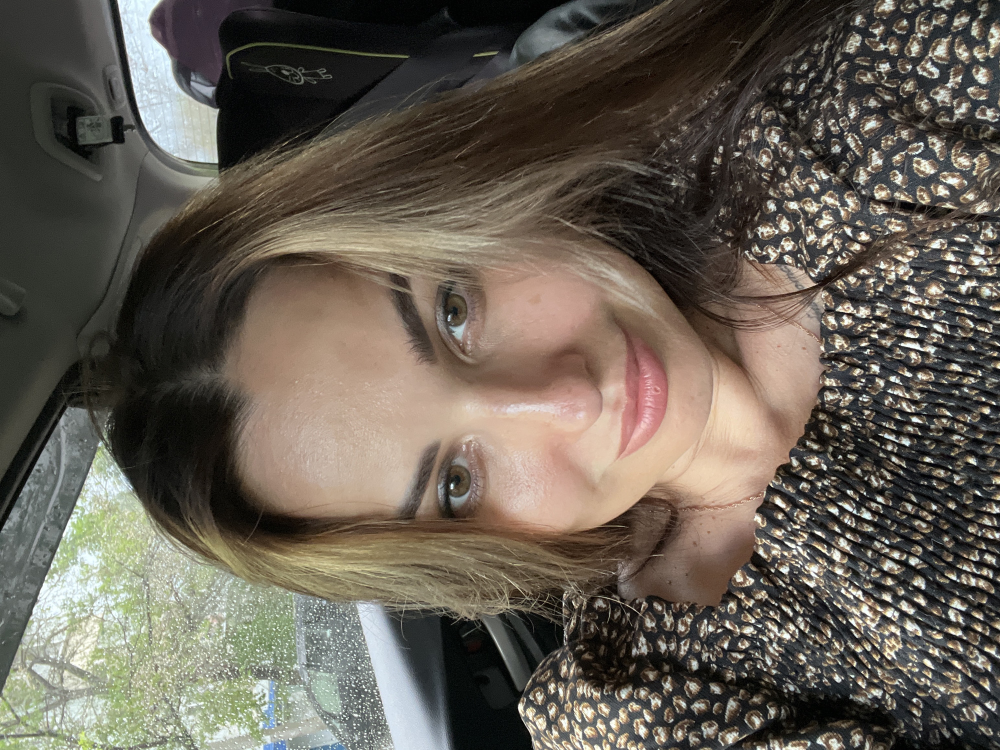 A woman with long, brown and blonde streaked hair is taking a selfie inside a car. She is wearing a black and brown leopard print top. Her hair is partially covering her face. She has light brown eyes and is smiling. The background shows a rainy day through the car window, with trees and some buildings visible. There is a child car seat in the back of the car. The car appears to be parked on a street in an urban area.