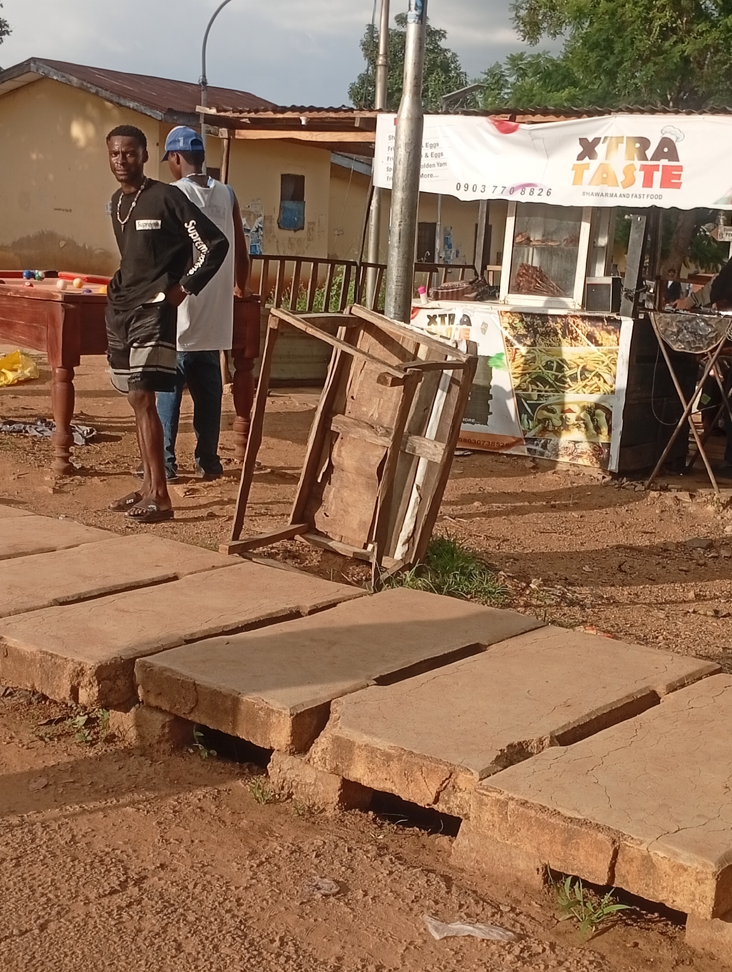 This media file captures an outdoor scene in Rafin Par, Nigeria, likely during the daytime given the clear lighting and shadows. The primary subjects are two young men standing near a wooden pool table on the left and a food stand on the right. The man in the foreground is wearing a black long-sleeved shirt with "Supreme" printed on it, black shorts, and sandals. He is looking towards the camera with a neutral expression. The second man, partially visible, is wearing a white shirt and a blue baseball cap, with his back to the camera.
The food stand, branded "XTRA TASTE Shawarma and Fast Food," is a prominent feature. A banner with the logo and a phone number (0903 770 8826) is displayed. The stand appears to be serving food, with skewers of meat visible inside a display case. The surrounding environment is dusty and appears to be an open area, possibly a market or street. In the foreground, there are concrete slabs, some of which appear broken or uneven, suggesting a less-than-pristine setting. A discarded wooden frame lies on its side, adding to the unpolished atmosphere. The background shows a building with a corrugated roof and some trees, indicating an urban or semi-urban setting. The overall impression is a snapshot of everyday life at a local eatery.