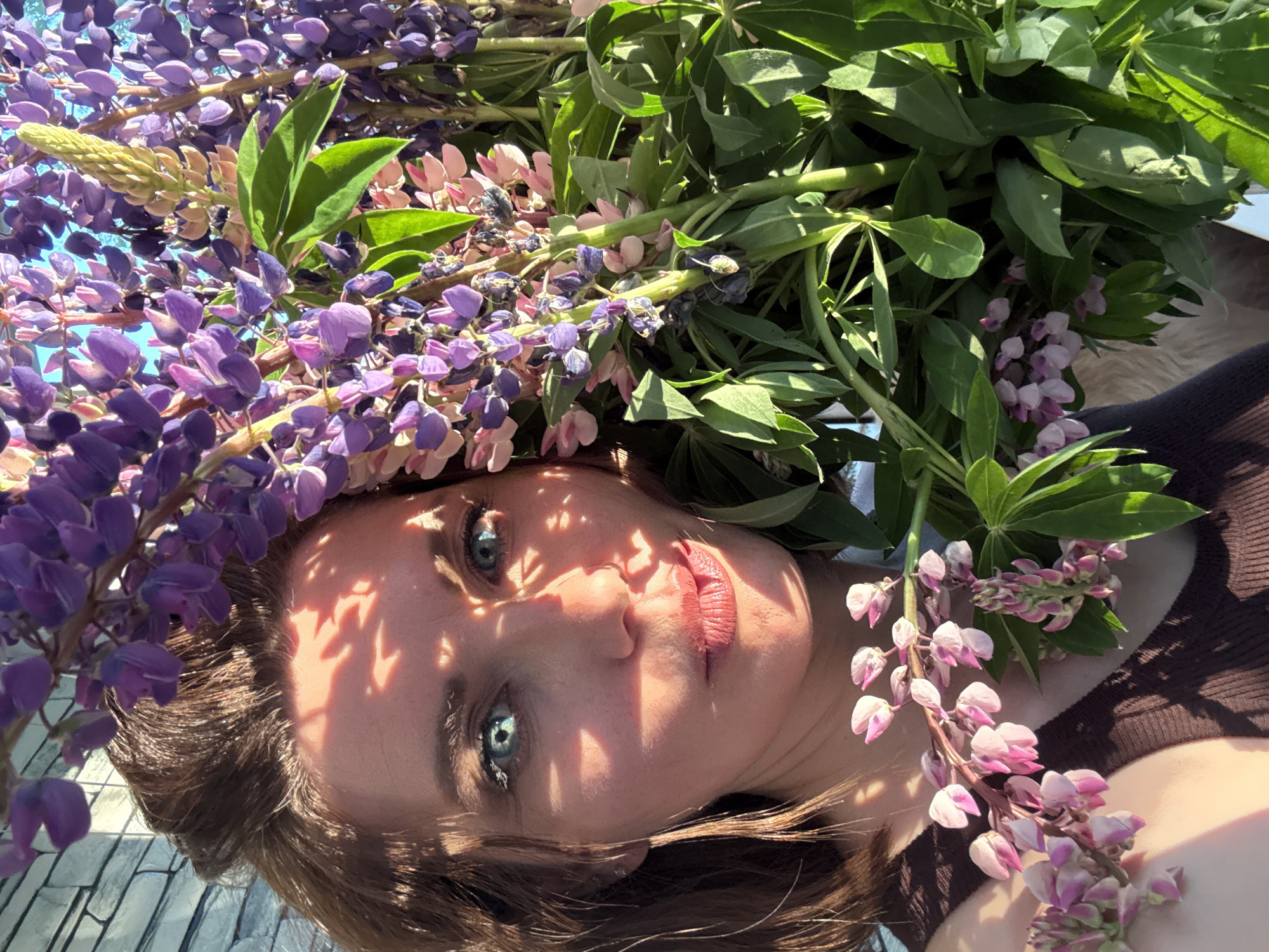 A medium shot captures a woman's face partially obscured by a large bouquet of lupine flowers. She has brown hair and striking blue-green eyes, with sunlight dappling her forehead and cheeks through the leaves and flowers. Her lips are slightly parted, and she is looking directly at the camera. She is wearing a dark brown ribbed tank top. The background is out of focus, but appears to be an outdoor setting with a hint of a brick wall on the left. The lighting suggests it is a sunny day, possibly late afternoon or early morning due to the warm tones. The flowers are a mix of purple and pink hues, with green foliage. The image does not provide any specific information that could identify the location within the city of Chaikovsky, Russia.