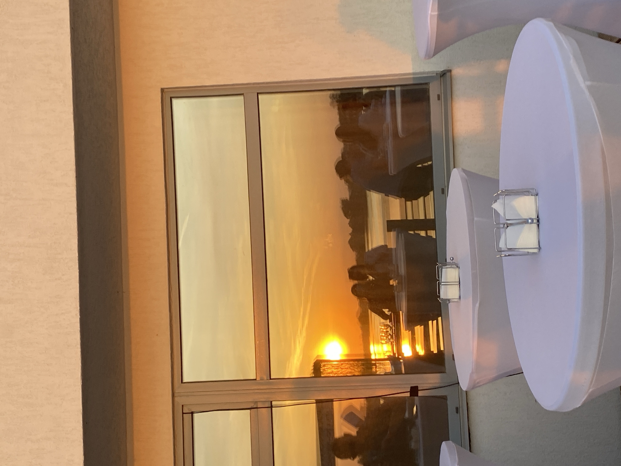 A sunlit room shows a window reflecting an outdoor sunset scene. People are reflected sitting at outdoor tables. There are also white-covered tables with small lanterns in the room. The location may be Aramil, Russia.