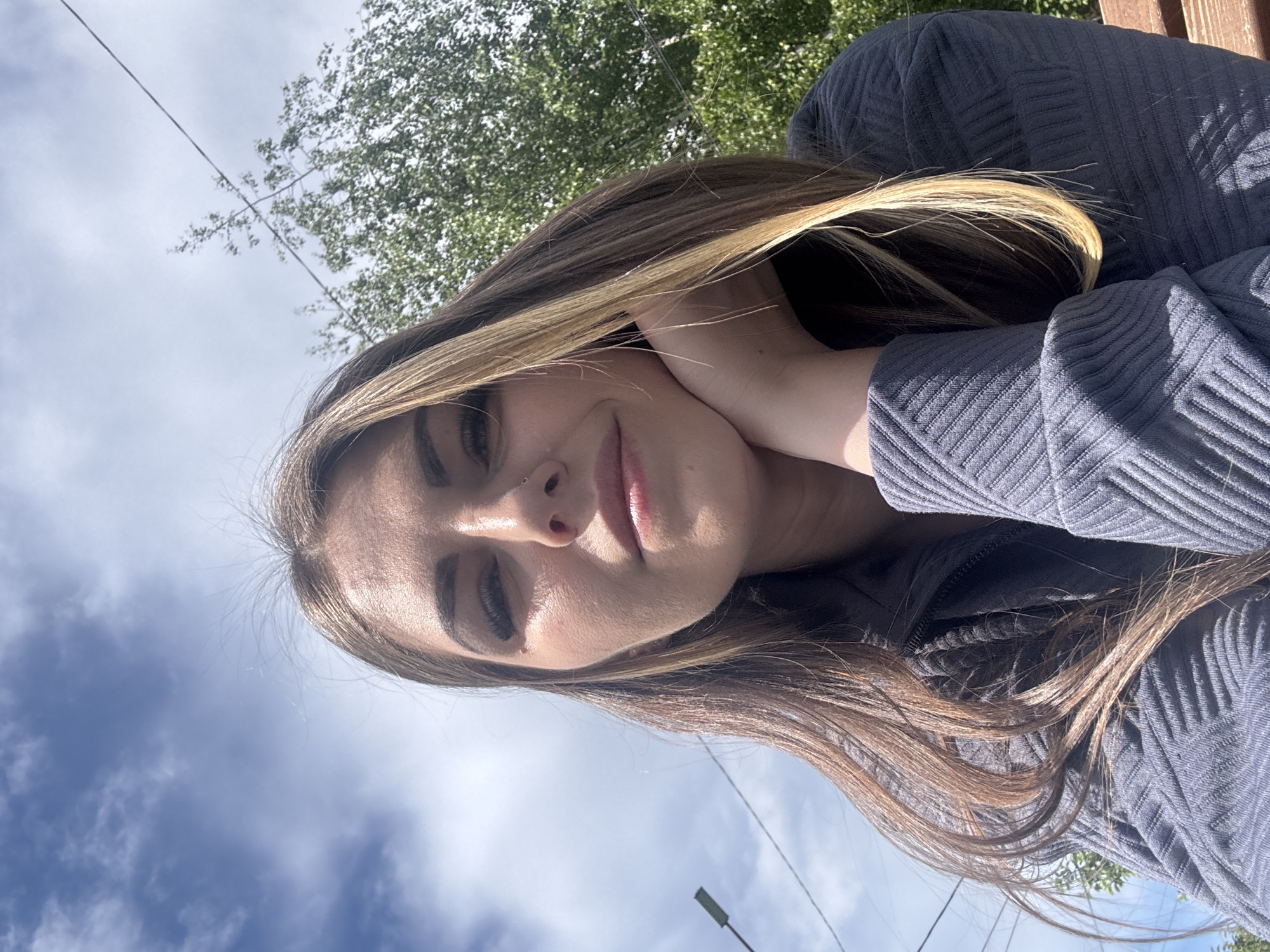 A young woman rests her face on her hand, sitting outside on a sunny day in what appears to be a park. The location is Shevlyakovo, Russia. Her light brown hair is streaked with blonde highlights. She is wearing a gray, textured, long-sleeved shirt or jacket. Overhead, there's a view of a blue sky with scattered white clouds and green tree foliage. A utility pole and wires are also visible.