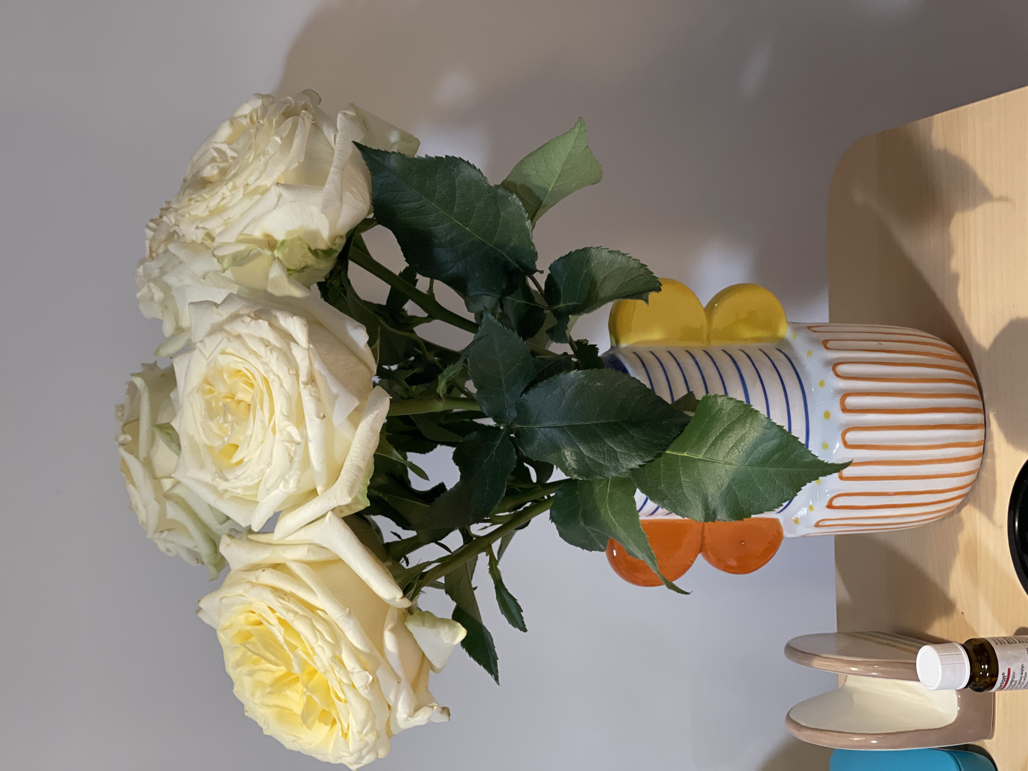 The image presents a still life composition featuring a vibrant bouquet of white and cream roses. The roses, with their full, intricate petals and dark green leaves, are arranged in a distinctive, colorful vase.
The vase is a prominent object, characterized by its playful design. It has vertical blue stripes on one side and a pattern of wavy, horizontal orange stripes on the other. Several rounded, bright yellow and orange shapes protrude from its upper section, adding to its unique aesthetic.
The background is a plain, light-colored wall, likely white or light grey, with soft shadows cast upon it, suggesting indoor lighting. To the right, a light wooden surface, possibly a shelf or tabletop, provides a resting place for the vase. On this wooden surface, a small, multi-tiered ceramic stand holds a tiny white bottle with a dropper cap. Faint text is visible on the bottle's label, including what appears to be "Naturals" and possibly "Eucalyptus."
The overall scene conveys a sense of calm and decorative elegance, typical of an indoor setting. No specific activity is taking place. The lighting is soft, creating subtle contrasts and highlights on the roses and vase.