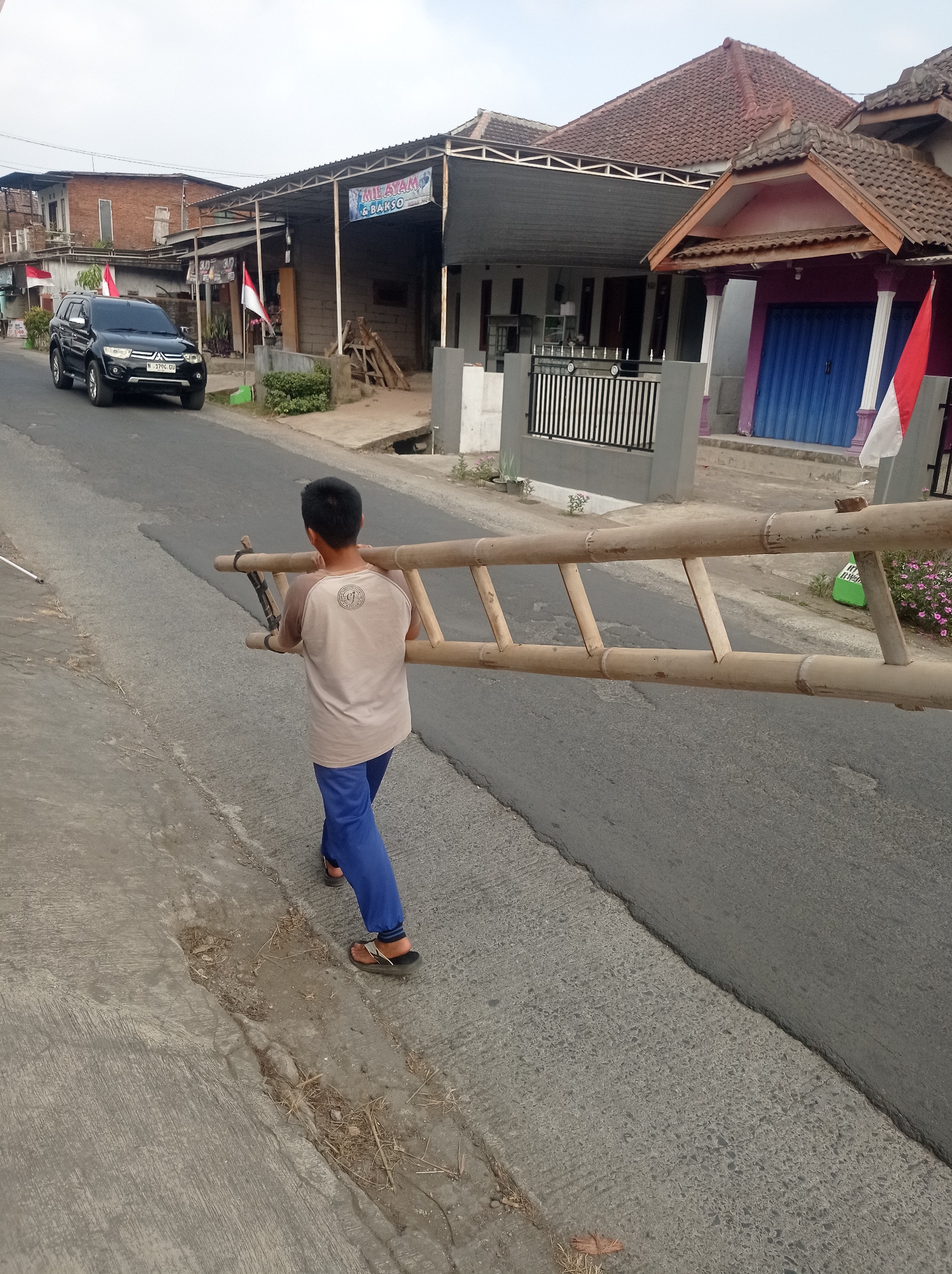 A young male, viewed from the rear, carries a long, makeshift bamboo ladder horizontally across his shoulders, walking on the right side of a paved road. He is wearing a light brown t-shirt with a circular graphic on the back and blue pants. He has dark hair and is wearing dark-colored flip-flops. The ladder extends from his left side to his right, with its ends reaching beyond the frame on both sides. The road surface is asphalt with some visible patches and cracks. To the left of the road is a concrete sidewalk with debris and dry grass.
In the background, on the right side of the road, are several buildings. One building has a reddish-brown tiled roof and a section with a dark gray overhang. There is a purple facade with blue roll-up doors and white columns. An Indonesian flag, red and white, is mounted on a pole to the right of this building. Another building behind it has a lighter gray facade and a black metal fence. Further back, a larger building with a dark gray metal roof structure is visible, and beyond that, a brick building with red tiles. A black SUV is parked on the left side of the road, facing away from the viewer, with an Indonesian flag attached to its windshield. A sign above a shop entrance reads "MIE AYAM & BAKSO". The sky is overcast. The location is Purwosekar, Indonesia.