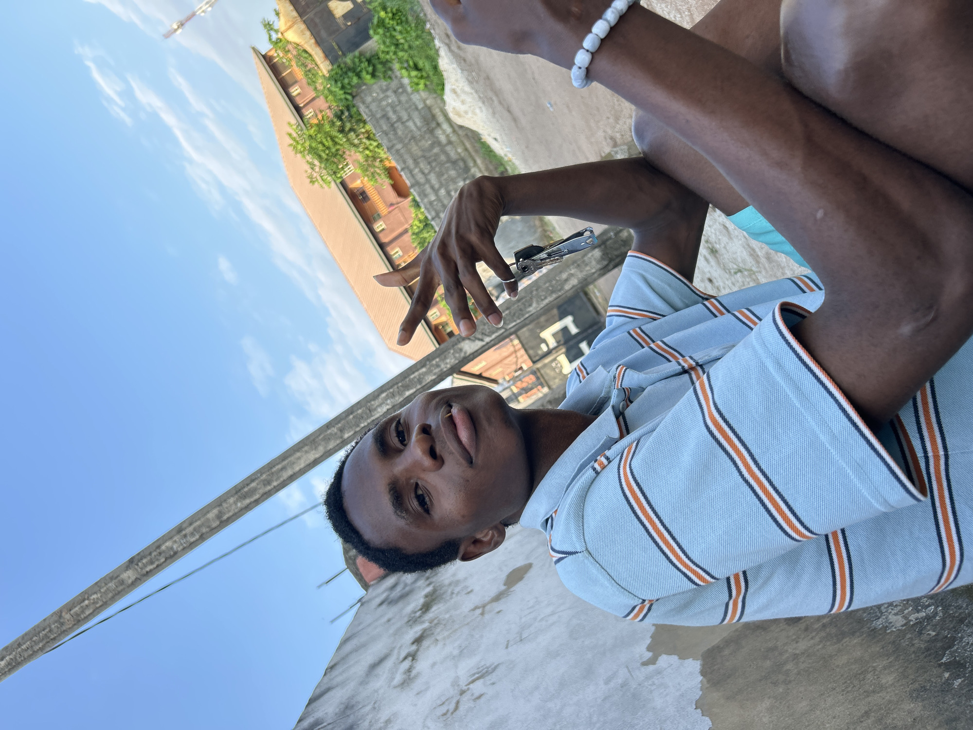 A young Black man is sitting outdoors, his body angled towards the camera. He is wearing a light blue collared shirt with thin orange and white stripes. His left arm is bent, resting on his lap, and his right arm is extended, holding a set of keys attached to a small silver multi-tool. He has a white beaded bracelet on his left wrist. He is looking directly at the camera with a neutral expression.
The background consists of a clear blue sky with scattered white clouds, and some buildings and greenery visible in the distance. A concrete structure, possibly a wall or rooftop, is in the foreground and beneath the man. The lighting suggests it is daytime, likely late morning or afternoon. The man appears relaxed, posing for the photograph. No visible text is present in the image. The scene is set in Ihiagwa, Nigeria.