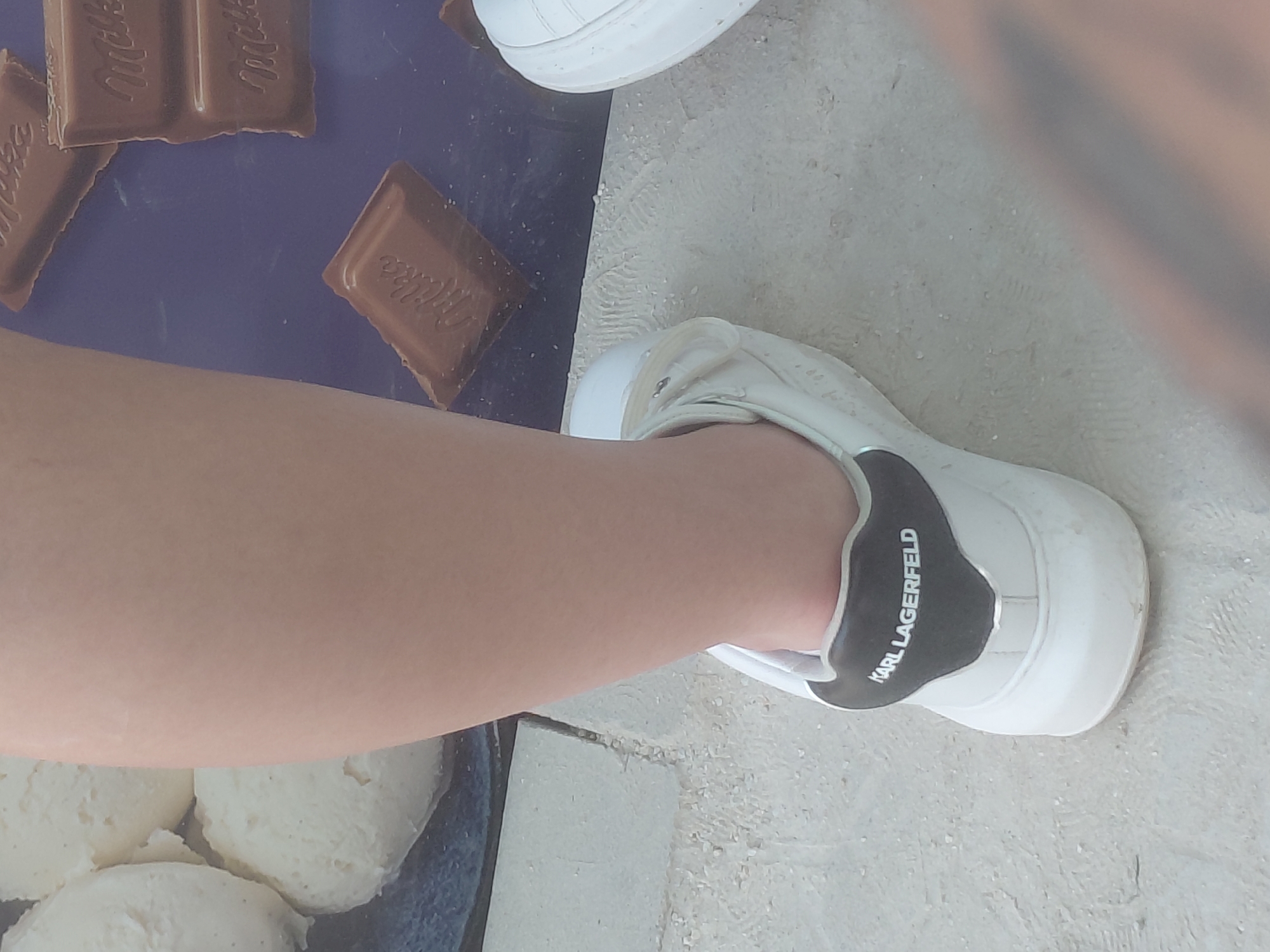 A high-angle forensic shot captures a person's lower right leg and foot, clad in a white Karl Lagerfeld branded sneaker with a black heel tab, standing on a light-colored, possibly tiled or paved, ground in Kostievo, Bulgaria. The sneaker exhibits minor dust accumulation. To the left of the leg, several scoops of a light-colored, creamy substance, resembling ice cream or mashed potatoes, are visible within a dark container. To the right of the leg, on a dark blue surface, multiple pieces of Milka milk chocolate, some broken, are scattered. A portion of another person's leg/foot is partially visible in the lower right corner, out of focus. The overall scene suggests a casual environment featuring food items.