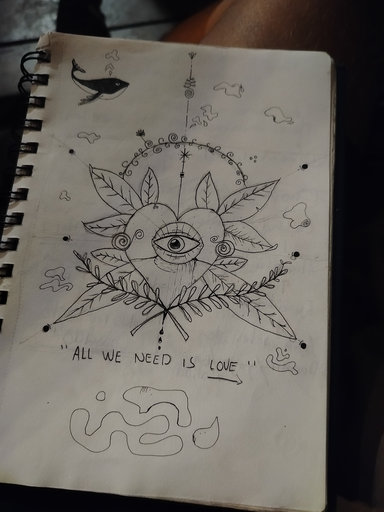 A spiral-bound sketchbook lies open, displaying a black ink drawing. The drawing features a central heart shape with an eye in the middle, surrounded by leaves, floral elements, and abstract shapes resembling clouds. Above the heart is a vertical line with stacked geometric forms. A small whale is depicted in the upper left corner. Below the heart, the phrase "ALL WE NEED IS LOVE" is written. The bottom of the page contains a meandering line drawing. The notebook appears to be resting on a dark surface. There is no evidence to indicate the location is in Playa del Carmen, Mexico.