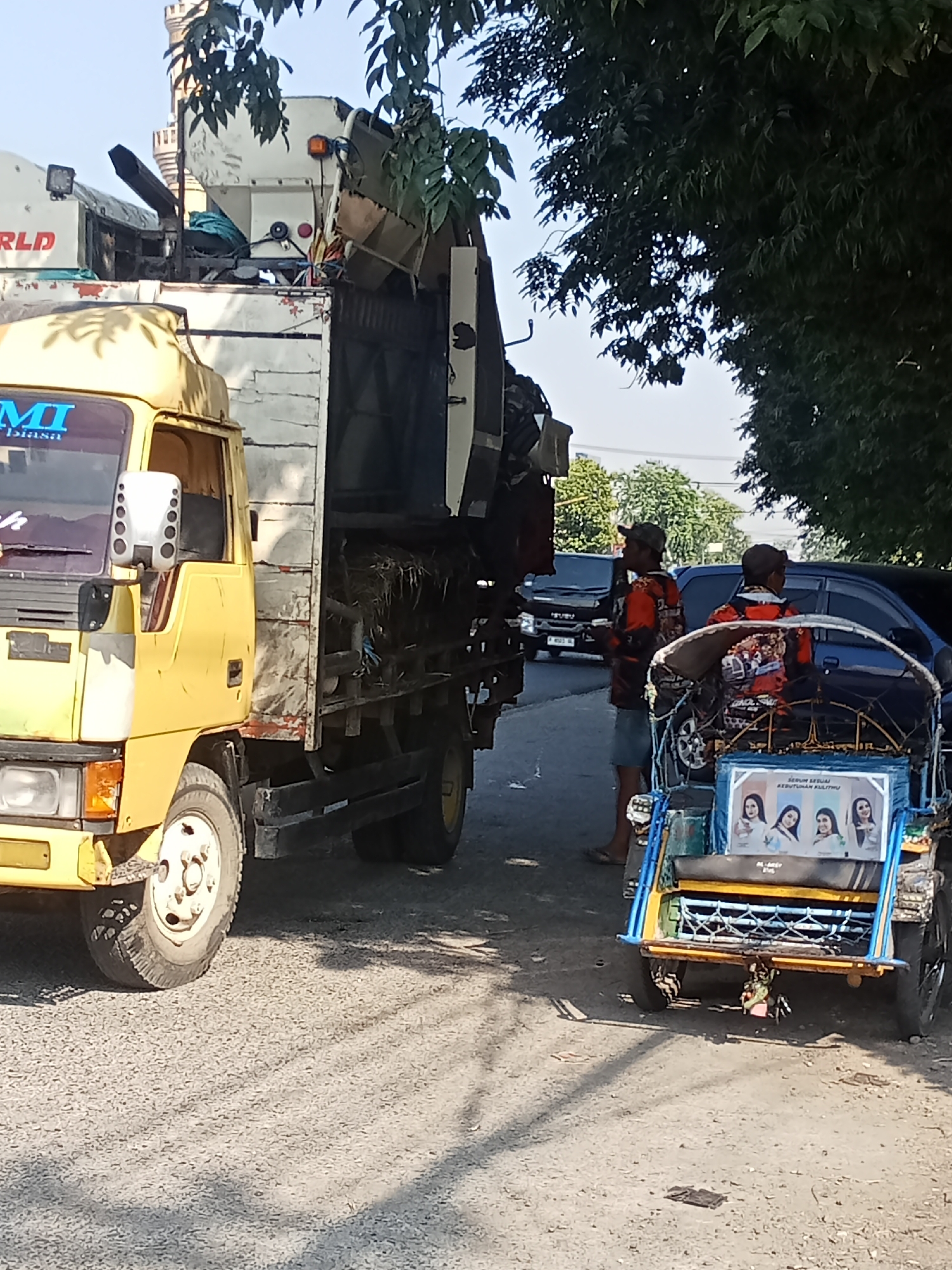 In Peterongan, Indonesia, two men wearing matching orange and black patterned shirts stand near a yellow truck and a blue and yellow motorized rickshaw. The man on the left holds a cup, while the man on the right looks toward the rickshaw. The rickshaw displays a poster with four women's faces and Indonesian text. The truck is loaded with various items, including what appears to be hay or dried foliage. In the background, a black SUV and other vehicles are visible, along with trees and buildings under a clear sky.