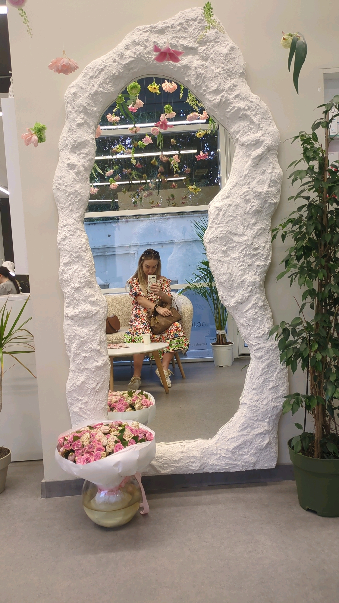 A woman with blonde hair and sunglasses is taking a selfie in a large, irregularly shaped mirror with a textured white frame. She is wearing a floral print dress and sitting on a cream-colored sofa. A brown handbag rests on her lap, and a white cup is on a small white table next to her. The mirror reflects a room decorated with numerous hanging artificial flowers in various shades of pink and green. In front of the mirror, on the floor, is a bouquet of pink and white roses in a clear glass vase. To the right of the mirror, a large potted green plant stands. The flooring appears to be a neutral gray concrete. The background visible through the mirror suggests an indoor space with large windows. This scene is not identifiable as being in Chișinău, Moldova.