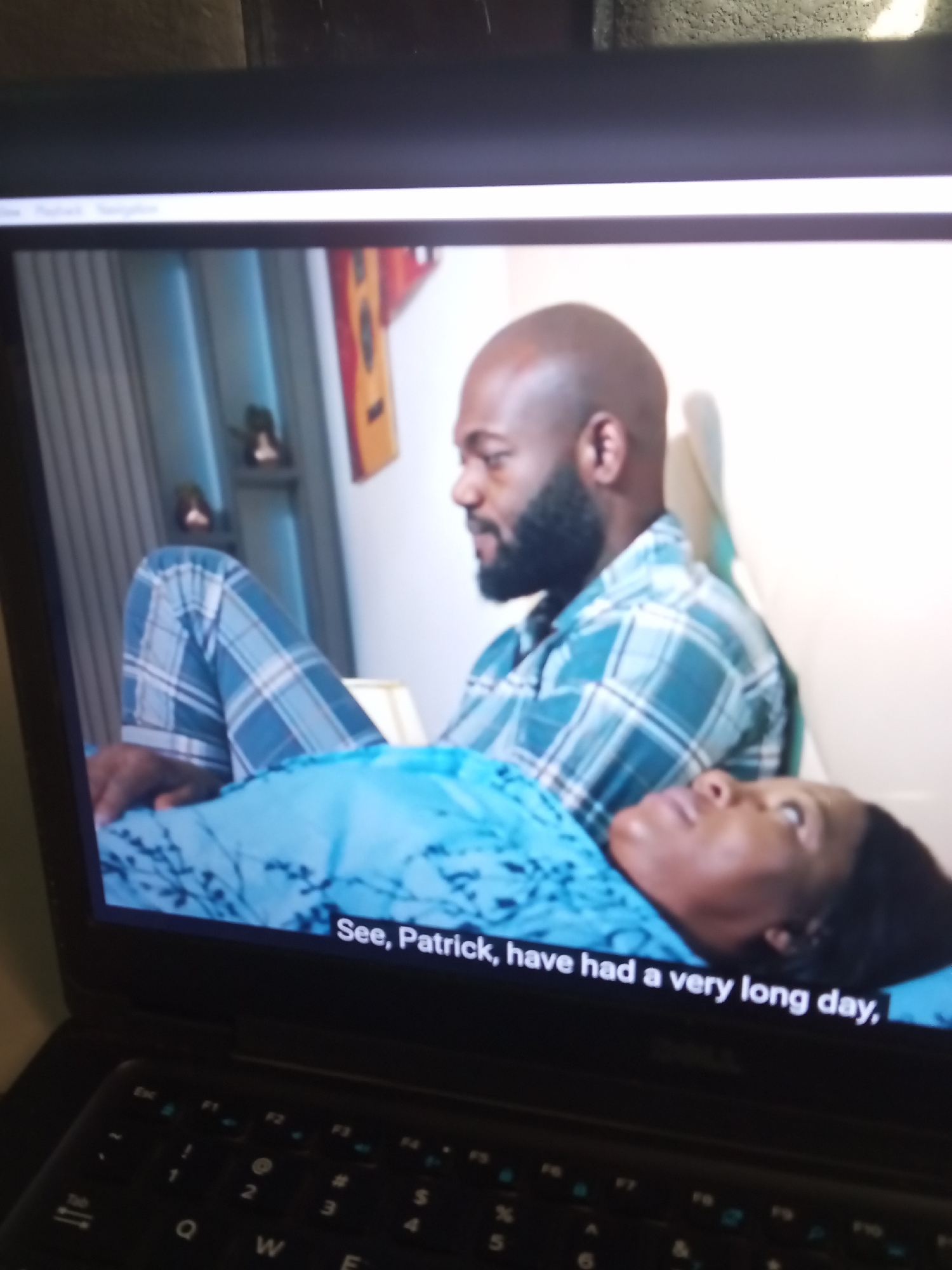 The image shows a close-up of a laptop screen displaying a scene from a video. On the screen, a man with a beard is lying down, wearing a blue and white plaid shirt and pajama pants. He is looking at a woman who is also lying down next to him, wearing a blue patterned top. A caption at the bottom of the screen reads, "See, Patrick, have had a very long day." The laptop keyboard is visible in the foreground, slightly out of focus. The background of the video scene appears to be a room with striped curtains and some decorative items on shelves.