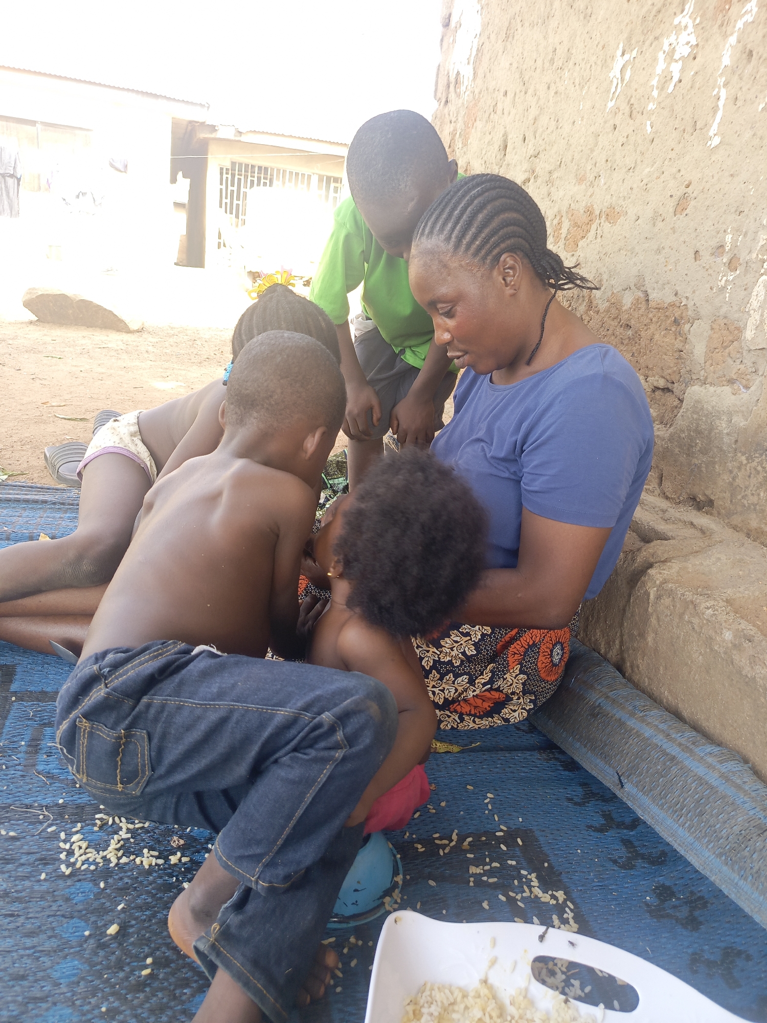 A woman sits on a blue mat beside a weathered wall in Akwanga, Nigeria, with several children gathered around her. She wears a blue short-sleeved shirt and a patterned skirt and appears to be tending to a young child. Three other children surround them; one boy is shirtless and wears jeans, another wears a green shirt, and the third wears a diaper or underwear. A white plastic container filled with food sits on the mat near spilled grains. The location appears to be outdoors, with buildings visible in the background.