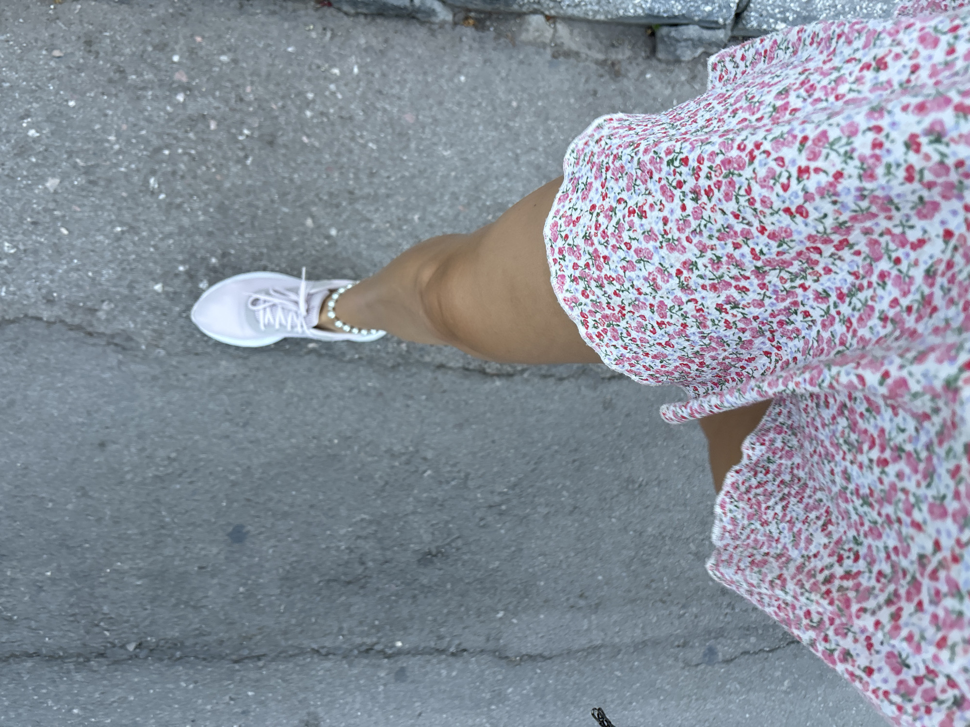 The image shows a first-person perspective of someone walking on a paved surface in Budva, Montenegro. The subject is wearing a short, floral print dress with pink and white flowers and a pair of light pink athletic shoes. A beaded bracelet is visible on their ankle. The ground is asphalt with some small stones and a faint line running down the left side, suggesting a crack or seam. To the right, the edge of the pavement meets a rough, gray textured surface, possibly a curb or wall. The lighting suggests it is daytime.