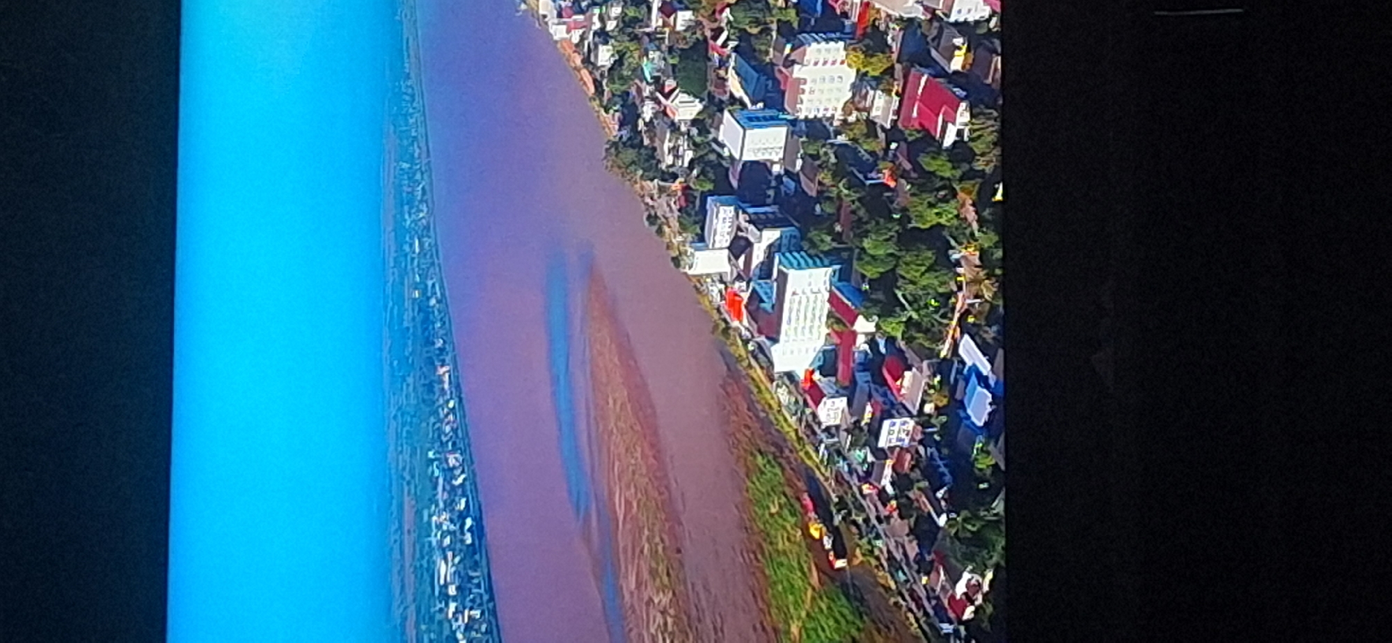The image depicts an aerial view of a city situated along a wide, murky river. The city appears to be densely populated with buildings of varying heights and styles, many of which have red tiled roofs. Lush green trees are interspersed among the buildings. The river is a prominent feature, with a sandy bank visible in the foreground. In the background, the river stretches towards a hazy horizon, where a distant cityscape or more land is suggested. The sky is a clear, bright blue. The lower part of the image is dominated by a black frame, indicating it is likely a photograph of a screen. The provided information about Bila Tserkva, Ukraine is not visually verifiable from the image itself.